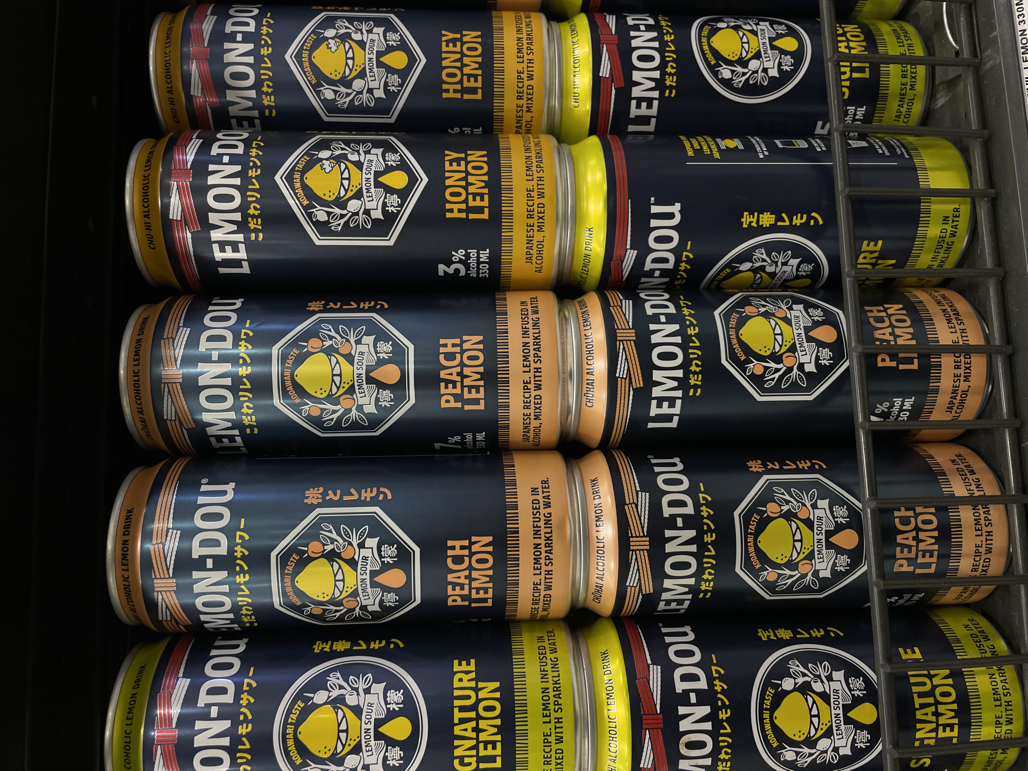 This is a close-up, eye-level shot of a retail display of canned alcoholic beverages, specifically "LEMON-DOU" chuhai lemon drinks, on a shelf in a store. The cans are arranged in multiple rows and columns, with some cans stacked horizontally on top of others. The lighting appears to be artificial, likely from store fluorescents, creating a slightly cool tone. The background is dark, suggesting a commercial setting like a convenience store or supermarket aisle. The cans themselves feature a dark blue label with prominent white lettering for the brand name "LEMON-DOU" and Japanese text below. Various flavors are visible, including "SIGNATURE LEMON," "PEACH LEMON," and "HONEY LEMON," each with distinct color accents on the label, such as yellow for signature lemon, orange for peach lemon, and a gold-yellow for honey lemon. The lower portion of some cans has a bright yellow or orange base with black vertical lines. There are no people visible in the frame. The overall impression is of a well-stocked retail display. The prompt requests the location be specified as Davao City, Philippines.