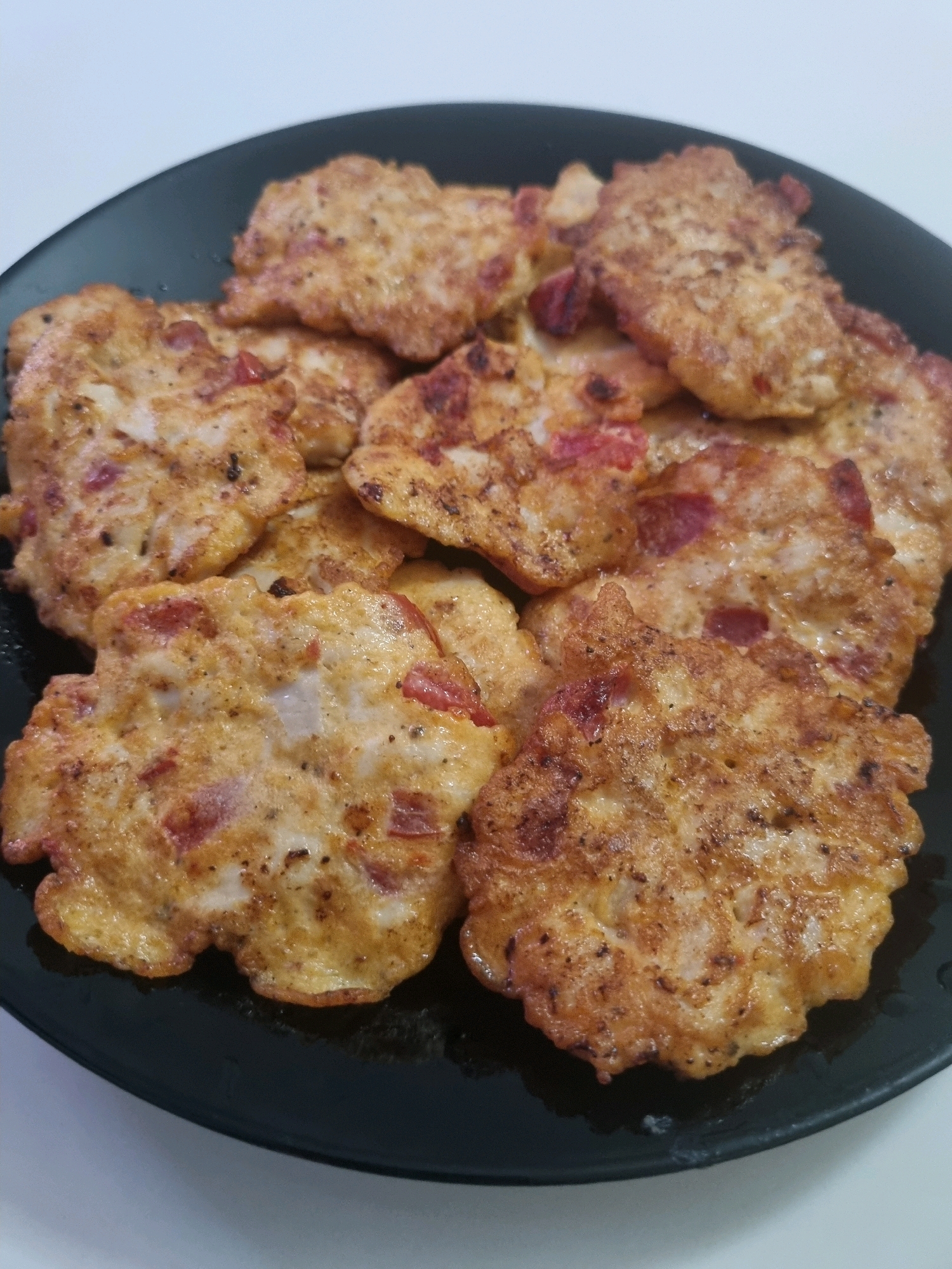A close-up shot shows a black plate filled with several round, golden-brown fritters. The fritters appear to be made of a mixture containing chopped red ingredients, possibly ham or vegetables, mixed with a light-colored batter, and have been pan-fried to a crispy texture. The lighting highlights the uneven surfaces and some darker brown spots on the fritters, indicating they are well-cooked. The plate is set against a plain white background, with a slight shadow suggesting the light source is from above and slightly to the left. There are no people visible in the image, nor any identifiable landmarks or context that would place it in the city of Bashkia Durrës, Albania.