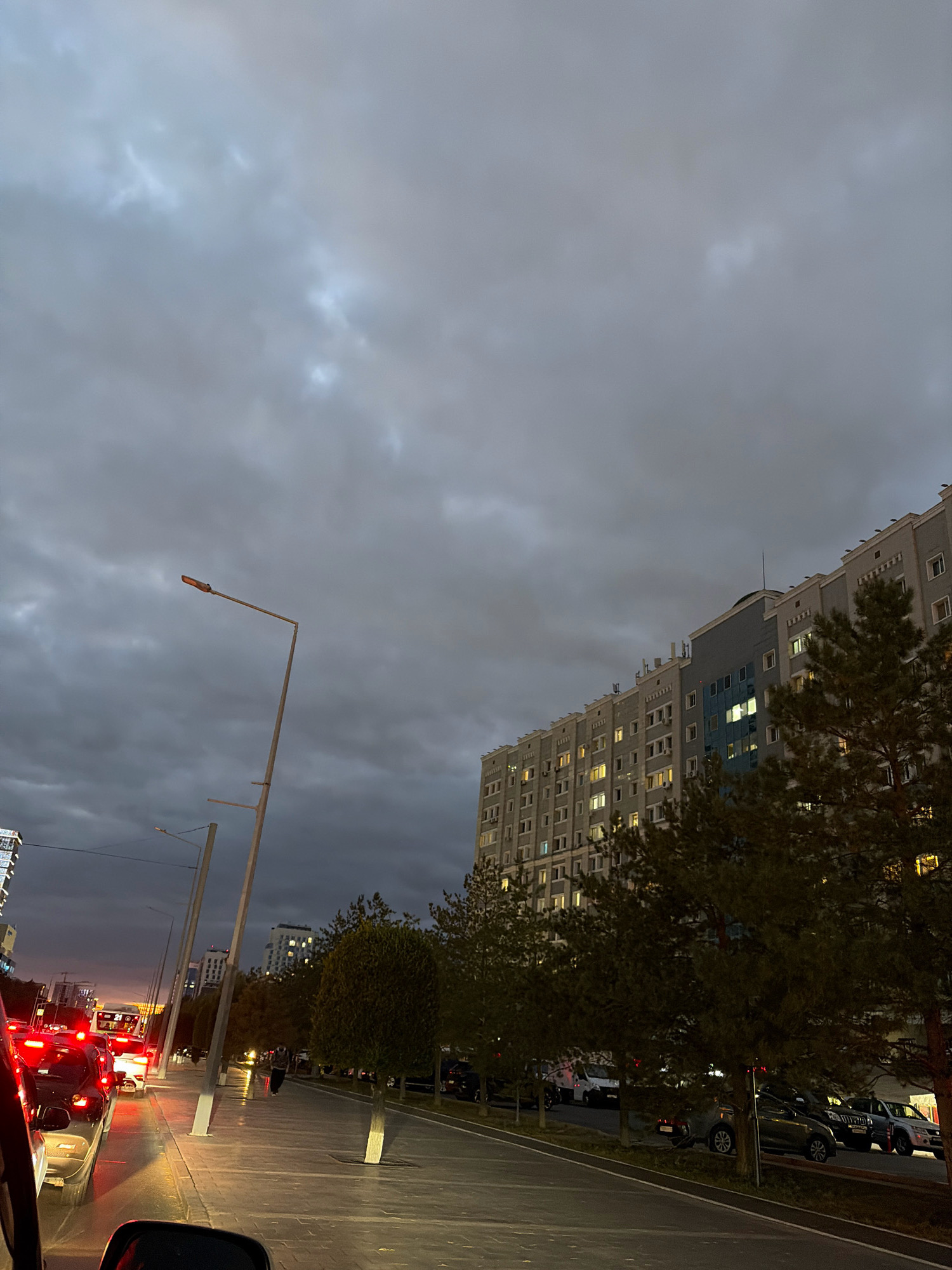 The image captures a street scene in Astana, Kazakhstan, during what appears to be dusk or twilight under a heavily clouded sky. The foreground is dominated by the interior of a vehicle, with the edge of a rearview mirror visible on the right. To the left, a line of vehicles is stopped in traffic, their red taillights illuminated, suggesting a congested urban environment.
Beyond the traffic, a wide sidewalk lines the street, illuminated by streetlights and the lights from a multi-story building. The building itself is a prominent feature, a long, grey structure with numerous windows, many of which are lit, indicating occupancy. A few trees, some with sparse foliage, are planted along the sidewalk, and a single pedestrian is visible walking on the path.
In the background, more buildings of varying heights are visible, some with illuminated windows. The sky is a uniform gray, with darker cloud formations creating a somber atmosphere. The overall impression is of a typical urban evening in Astana, with the city's infrastructure and daily life unfolding under the dimming light.