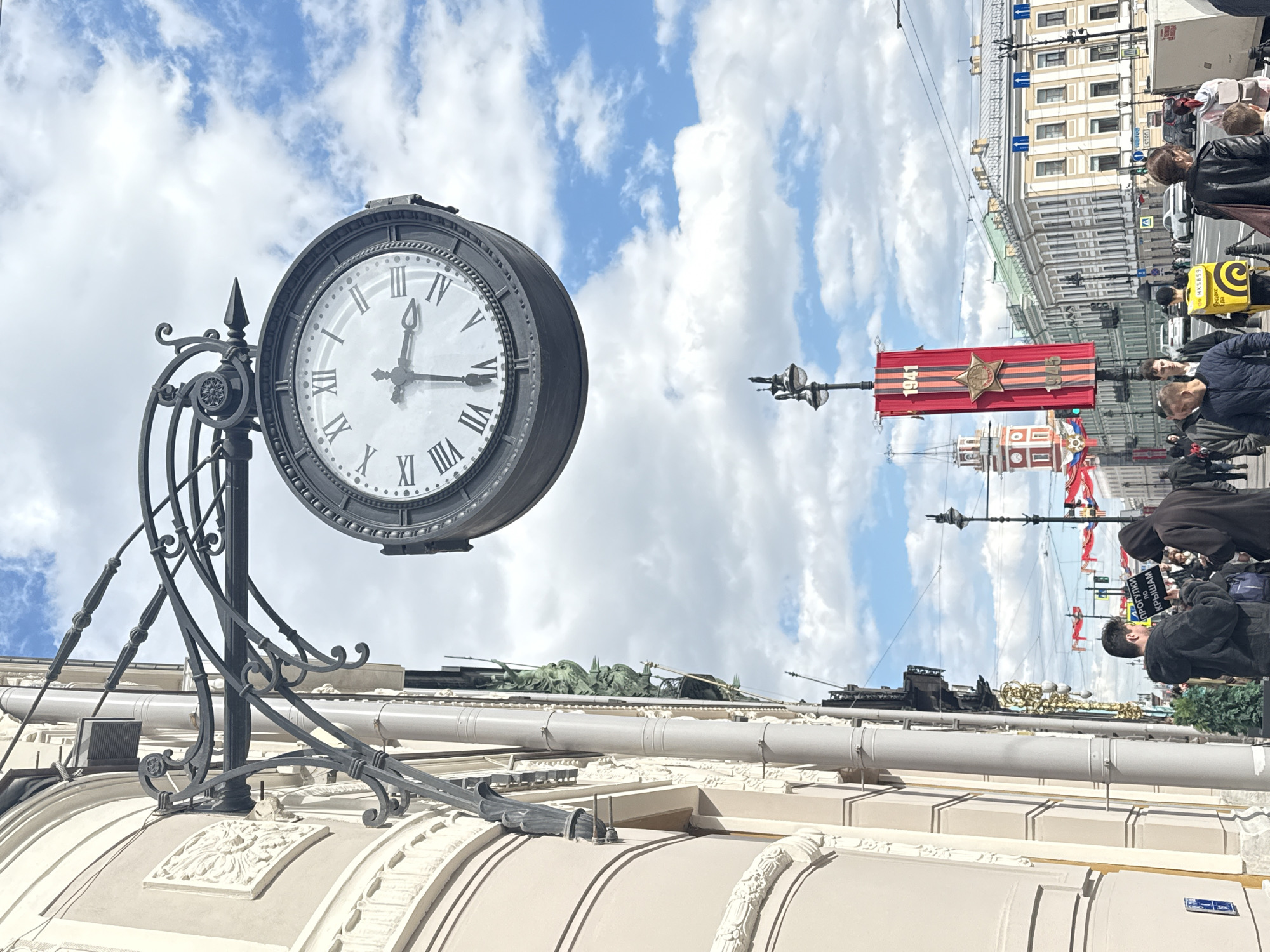 The image shows a street scene in Saint Petersburg, Russia, likely along Nevsky Prospekt. In the foreground, an ornate, antique-style clock is attached to the facade of a building, displaying the time as approximately 10:34. Above the clock, a partly cloudy sky is visible. In the background, a crowd of people is gathered, some holding flags and banners, including a red banner adorned with a star and the years 1941-1945. Street lamps and building facades are visible among the crowd. The people seem to be observing a parade.