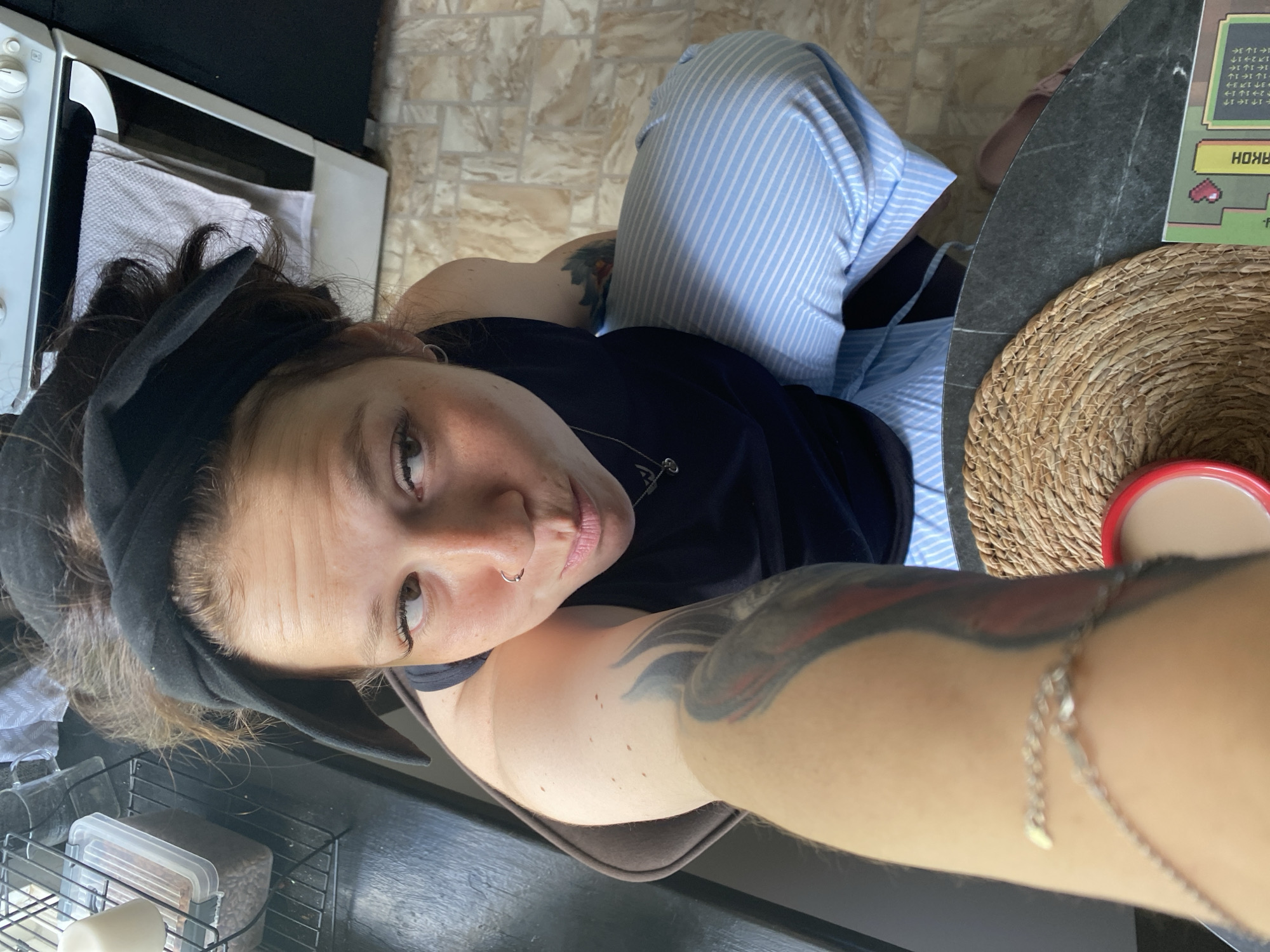 A woman with a black headband and a black tank top is sitting on a chair in what appears to be a kitchen. She is looking up and to the side with her lips pursed. She has a nose ring and a silver necklace with a heart pendant. She has tattoos on her left arm and shoulder. She is wearing light blue striped pajama pants. In the foreground, there is a woven placemat with a red mug on it. The floor is tiled with a beige and brown pattern. In the background, there is a white oven and a shelf with a plastic container of coffee beans. The location is the city of Chaikovsky, Russia.
