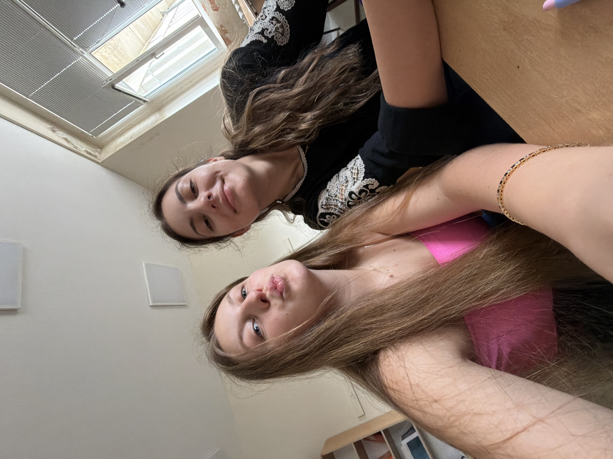 This media file captures two young women in an indoor setting in Burgas, Bulgaria. The image is rotated approximately 90 degrees clockwise, giving the impression that the subjects are looking down at the camera from above.
The woman closer to the camera (positioned in the lower-left due to rotation) has long, straight light brown hair, blue eyes, and is wearing a bright pink top. Her lips are slightly pursed in a "duck face" expression, and a delicate necklace with small heart-shaped pendants is visible on her neck. Her arm is partially visible, covered by her flowing hair.
Behind her (positioned in the upper-right), the second woman has long, wavy dark brown hair and a subtle, closed-mouth smile. She is dressed in a black long-sleeved top adorned with intricate white embroidered patterns on the sleeves and along the neckline. A gold-toned bracelet is visible on her right wrist, which rests on a light-colored wooden surface, possibly a table or desk.
The background elements include a window with horizontal blinds in the upper-left (if the image were rotated upright), through which an exterior building is partially visible, indicating daylight. White walls are prominent, featuring two square white panels, possibly speakers or minimalist wall-mounted lights. A wooden shelf unit is also partially visible in the bottom-right. The overall impression is casual and friendly. No text is visible in the image.