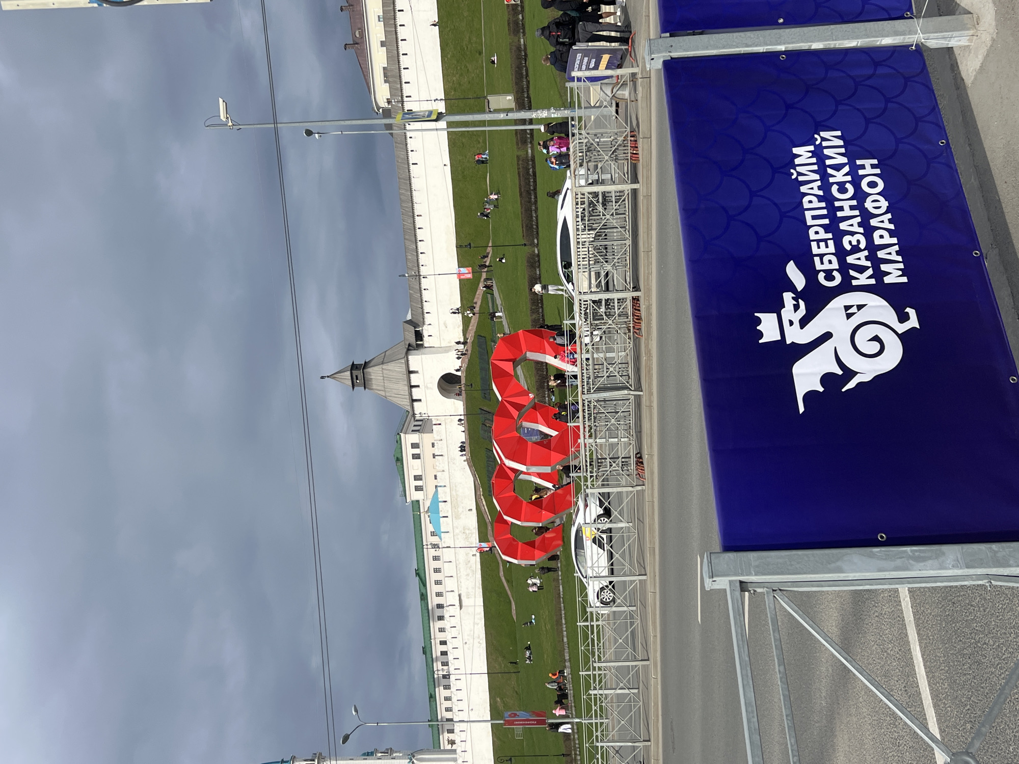 The image shows a view in Kazan, Russia, likely near the Kazan Kremlin. In the background are white-walled structures with pointed roofs, indicative of the Kremlin's architecture. In the foreground, there's a blue banner with white text and a dragon logo, possibly for a marathon event, and metal barriers lining a street. People are seen on a grassy area adjacent to the buildings. There's also what appears to be a red, looping sculpture or art installation. The sky is overcast.