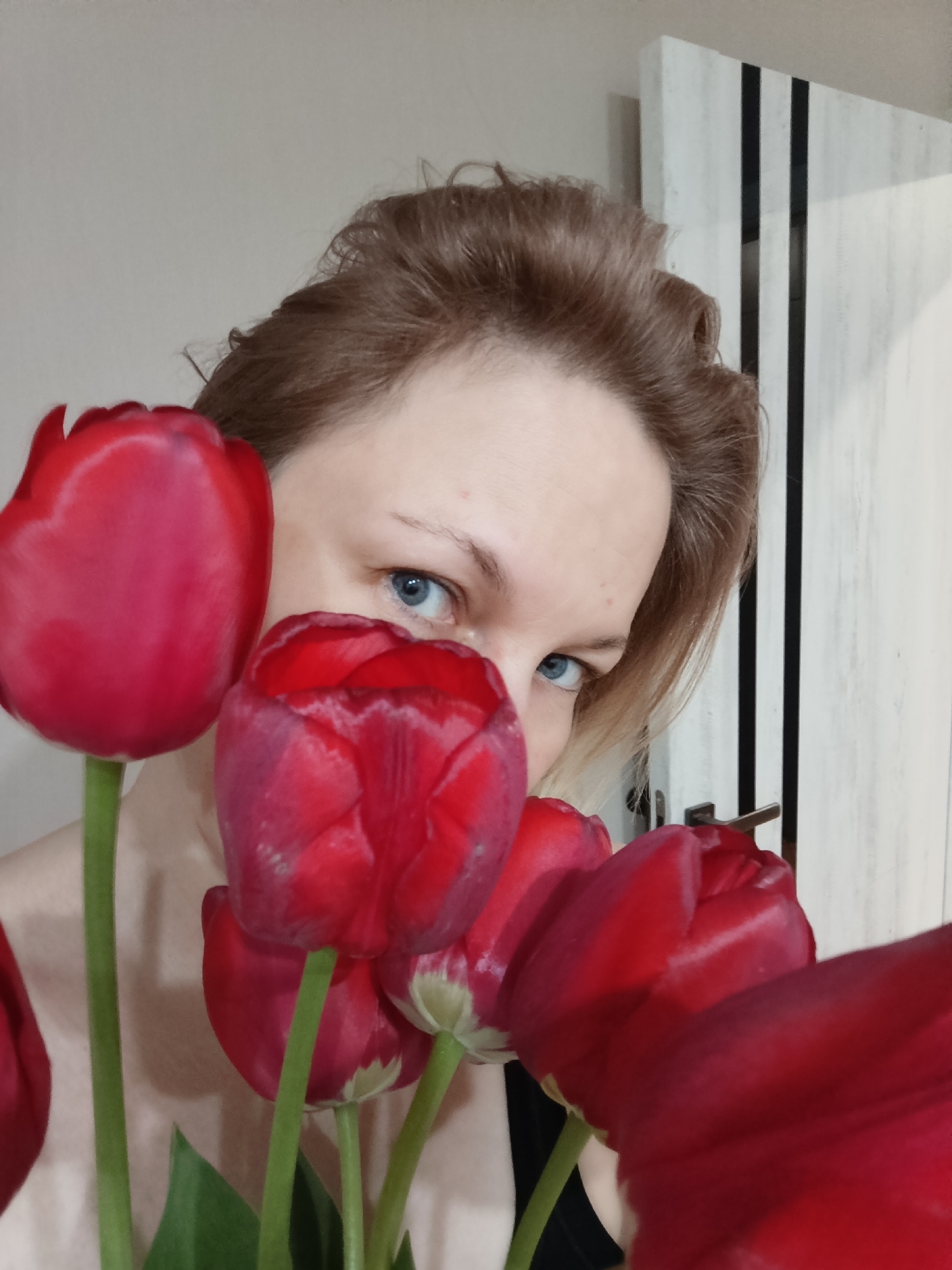 A woman with light skin and blue eyes is holding a bouquet of red tulips. The tulips are in the foreground, partially obscuring her face. Her hair is light brown and styled upwards. Behind her is a white door with black vertical stripes and a silver door handle. The location is suggested to be Марковский, Russia.