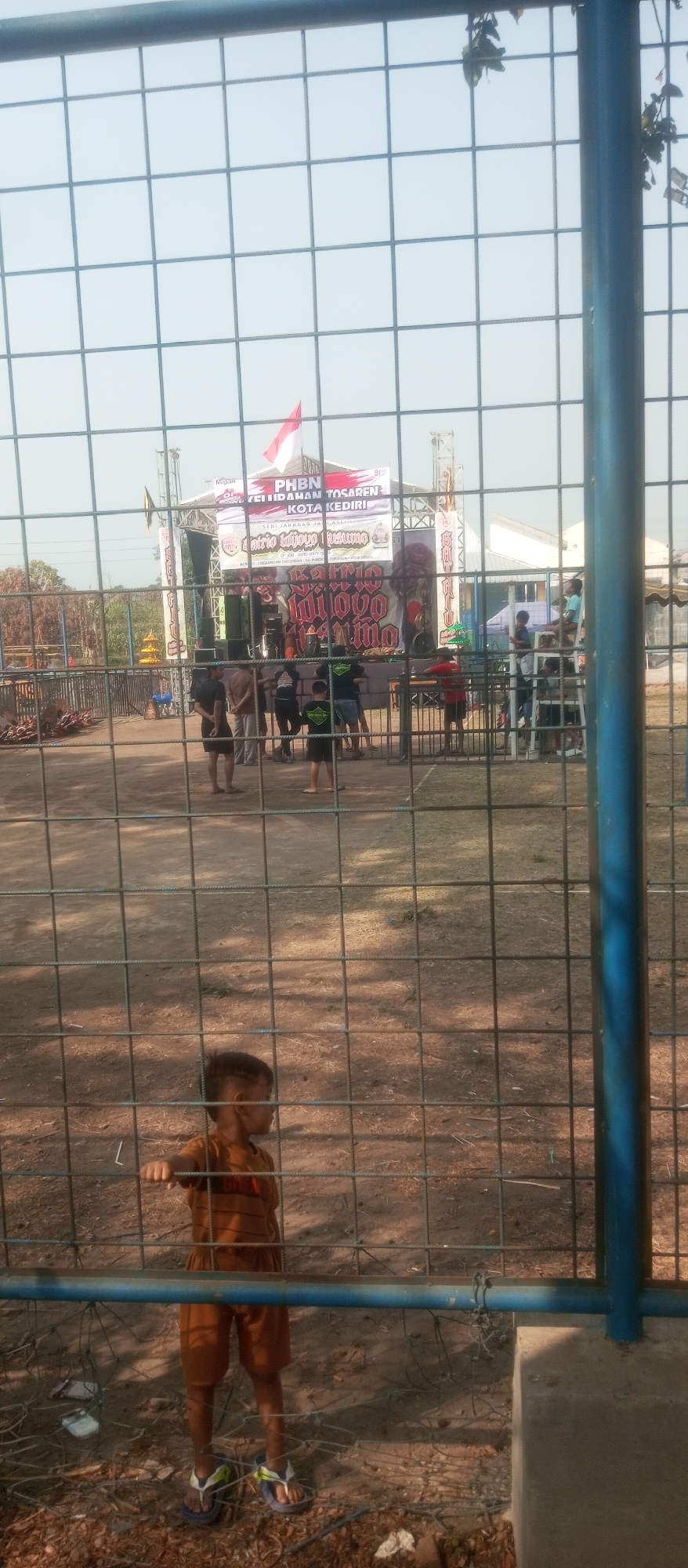 The image shows an outdoor event with a stage in the background, a fence in the foreground, and a young child standing near the fence. The stage has a banner with Indonesian writing and a red and white flag. Several people are visible around the stage, some of whom appear to be setting up equipment. The child in the foreground is wearing a brown striped shirt and brown shorts, with flip-flops on their feet. The ground is covered with dirt and some scattered debris. The overall scene suggests a public gathering or performance taking place in Kediri City, Indonesia.