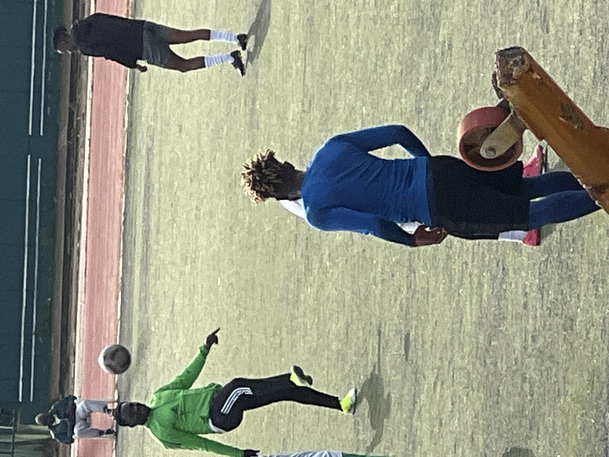 This image captures a moment on a dusty, outdoor sports field in the New Stadium area of Nigeria. The field itself appears to be a green, worn surface, possibly a soccer pitch, with a red track running parallel to the background. In the foreground, a person wearing a bright green long-sleeved jacket and black pants with white stripes is in mid-action, kicking a soccer ball. Their left leg is extended, and their right foot is poised to strike. The soccer ball is in the air.
Further back, another individual is dressed in a blue long-sleeved shirt and black shorts, facing away from the camera. To the left, a person is seated on a white folding chair, wearing a dark jacket and light-colored pants, observing the scene. Another figure in a dark jacket and shorts stands on the field to the right. The background features a dark green barrier or wall with horizontal white lines. A rusty orange-colored piece of equipment with a red roller is prominently positioned in the lower right corner, partially obscuring the field. The lighting suggests it is daytime.