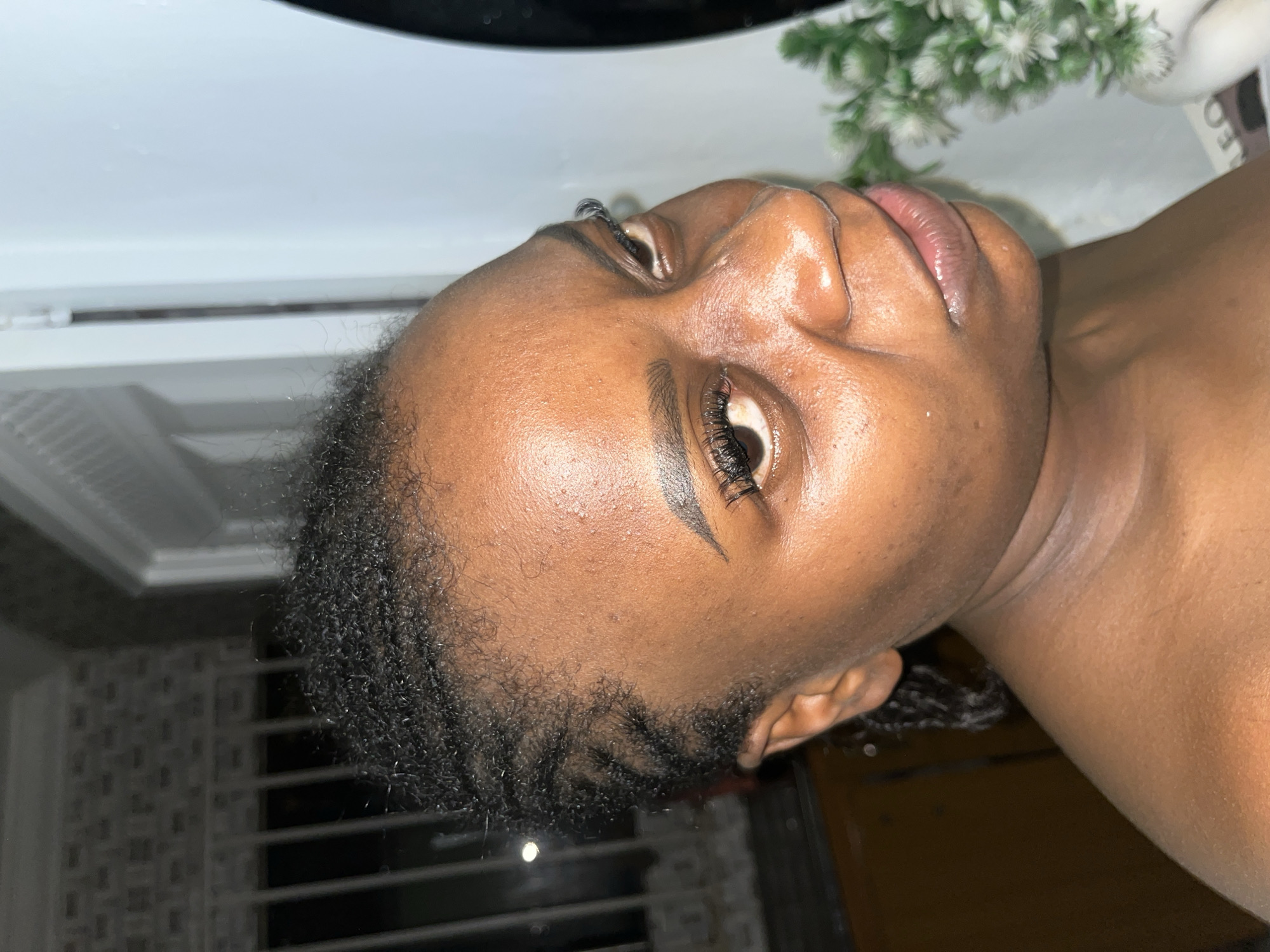 A medium close-up, side-profile shot captures a Black woman with braided hair, looking towards the right of the frame. She is positioned in an indoor setting, with a blurred background that includes what appears to be a white door and a dimly lit area with vertical bars. The woman's face is illuminated, highlighting her skin tone and facial features. She is wearing what appears to be false eyelashes. To the right of the frame, a portion of a white vase containing a green, leafy plant is visible. The lighting suggests an artificial light source. The scene is devoid of any explicit indicators of a specific city or location within Abuja, Nigeria.