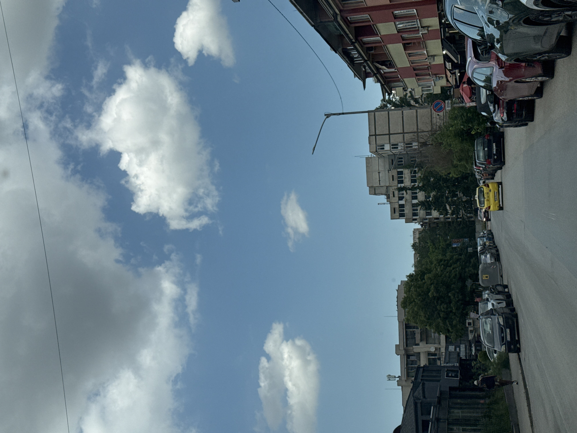 The image captures an urban street scene on a bright, clear day, likely in Sofia, Bulgaria. The sky, filling the upper portion, is a vibrant blue with scattered white cumulus clouds. A thin, dark cable or power line traverses the top left.
Along the right side of the image, a paved street is lined with numerous parked cars of various models and colors, predominantly dark. A bright yellow small car stands out among them. License plates such as "CA 6280 XM" and "CB 0860 HT" are visible on some vehicles. Further down the street, a person dressed in a dark jacket and light head covering is walking on the sidewalk.
The street is flanked by buildings. On the immediate right, a multi-story building features a distinctive red and beige checkered facade. Further along, several grey, concrete-like multi-story buildings are visible, partially obscured by green trees and bushes that add foliage to the urban landscape. A black streetlight extends over the road. A "No Parking" sign (red circle with a blue background and diagonal line) is partially visible behind some vegetation.
The bright sunlight suggests it is daytime, likely mid-day. The scene depicts typical daily life in an urban environment.
