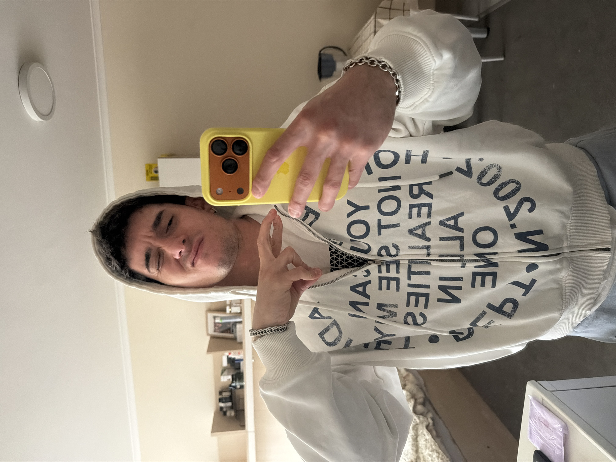 A man is taking a selfie in a room. He is wearing a white hoodie with black text, a silver bracelet, and holding a yellow smartphone. The room has a light beige wall, a white ceiling with a round light fixture, and a shelf with various items on it in the background. The image does not provide enough information to pinpoint the specific location in Sofia, Bulgaria.