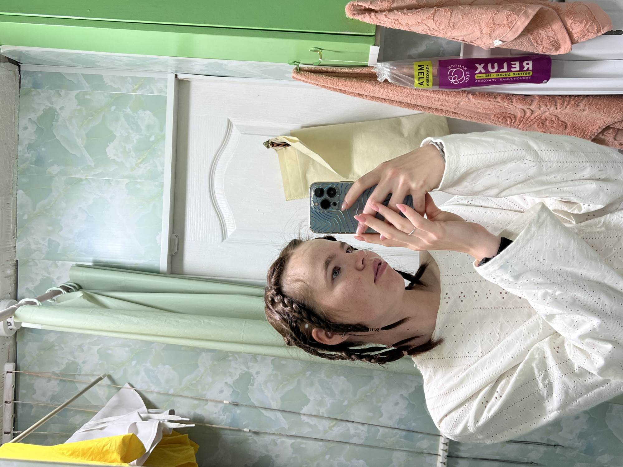 The image shows a young woman taking a selfie in what appears to be a bathroom. She has brown hair styled in braids and is wearing a white eyelet top. She is holding a blue smartphone in her hands, taking the photo.
The background includes a green cabinet, white marble-patterned walls, a white door, and a towel rack. There are peach-colored towels and a purple rectangular package on the shelf above the door. There are also clothes drying on the towel rack.
Based on the "RELAX" product visible, which has the words "Chaikovsky" and "Russia" on the packaging, the location is most likely in the city of Chaikovsky, Russia.
