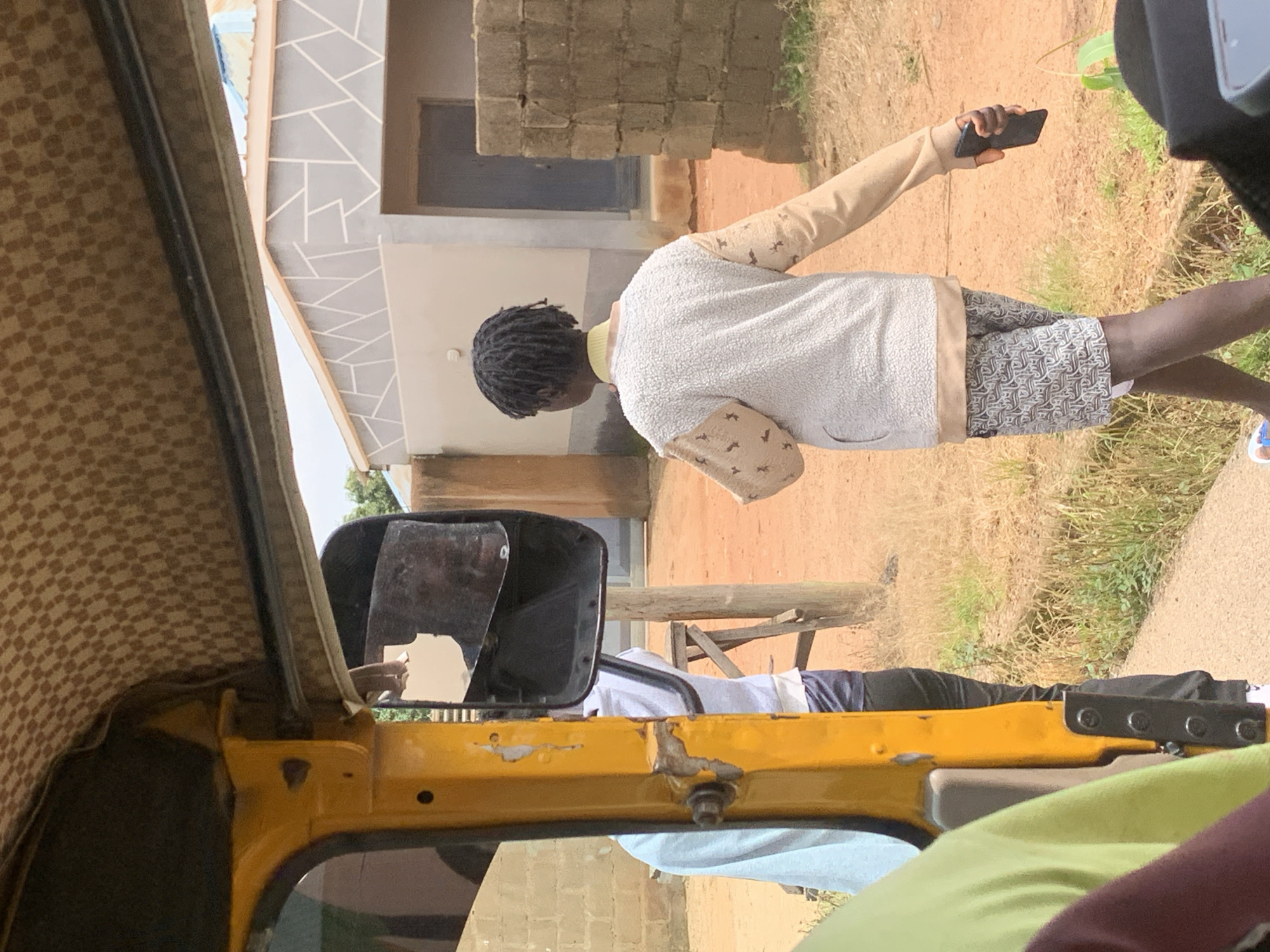 This media file depicts a scene in Jos, Nigeria, with a person standing outdoors. The person, seen from the back and side, is wearing a light-colored, textured sweater with a beige collar and decorative beige detailing on the sleeves. They are also wearing patterned shorts. The individual is holding a black smartphone in their right hand, appearing to take a picture or record a video.
The setting is outdoors, with elements of a building visible in the background, characterized by brickwork and a light-colored facade with geometric decorative lines. Dry grass and some vegetation suggest a somewhat arid environment, possibly during a dry season. The lighting indicates it is daytime, likely under a clear or partly cloudy sky, given the strong shadows.
The perspective suggests the photo is taken from within a vehicle, possibly a tricycle or auto-rickshaw, given the visible interior elements like a checkered fabric roof lining and a yellow painted metal frame of the vehicle. A side mirror of the vehicle is also prominent in the foreground, reflecting a portion of the scene and a hand. The overall mood is casual and observational, capturing a moment of everyday life. There is no visible text within the image.