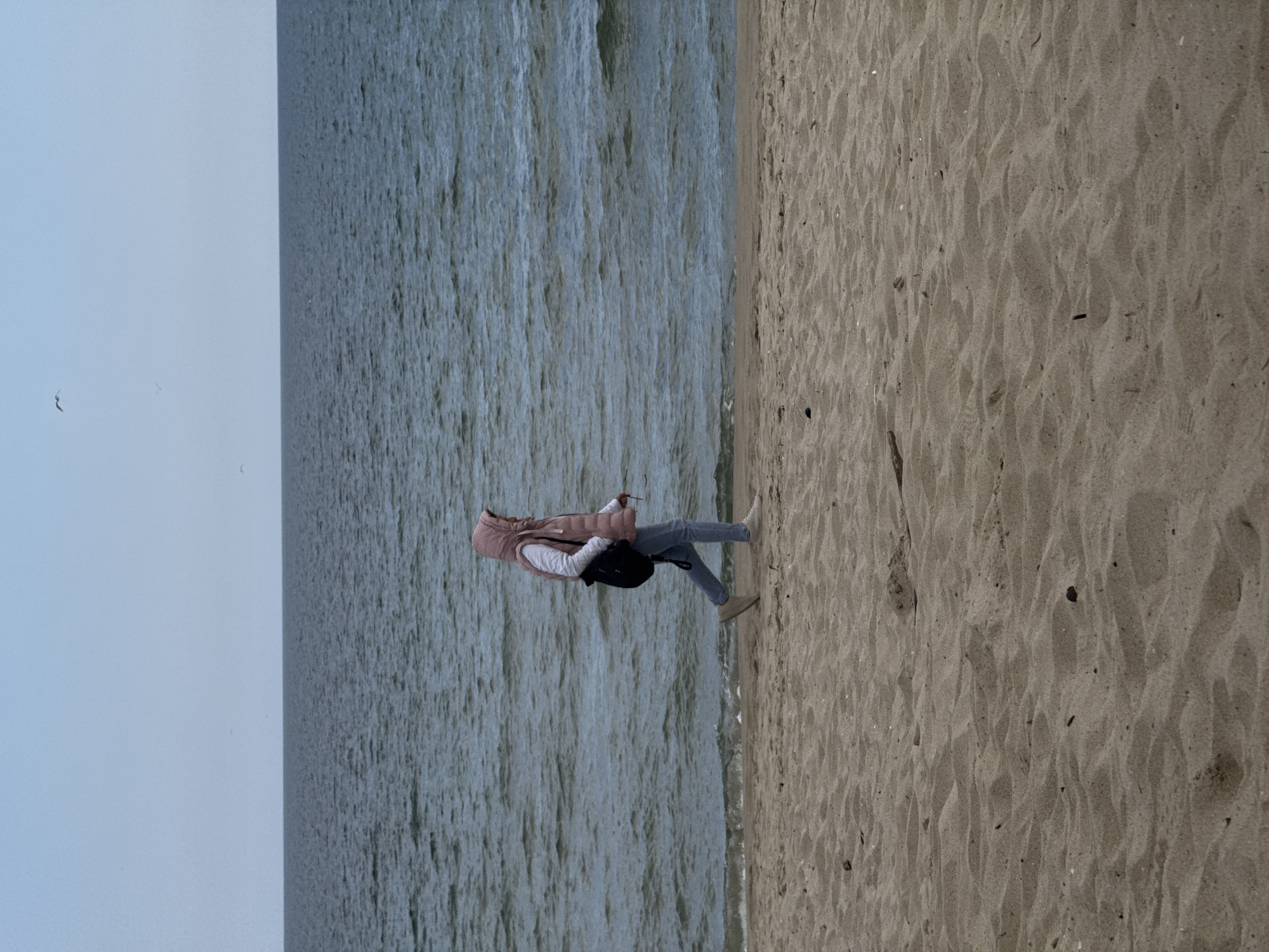 A person is walking on a sandy beach next to the water. The person is wearing a pink vest, a white top, jeans, and light-colored shoes, and carries a black bag. The location is likely a beach in Varna, Bulgaria, given the provided context. The sky is overcast.