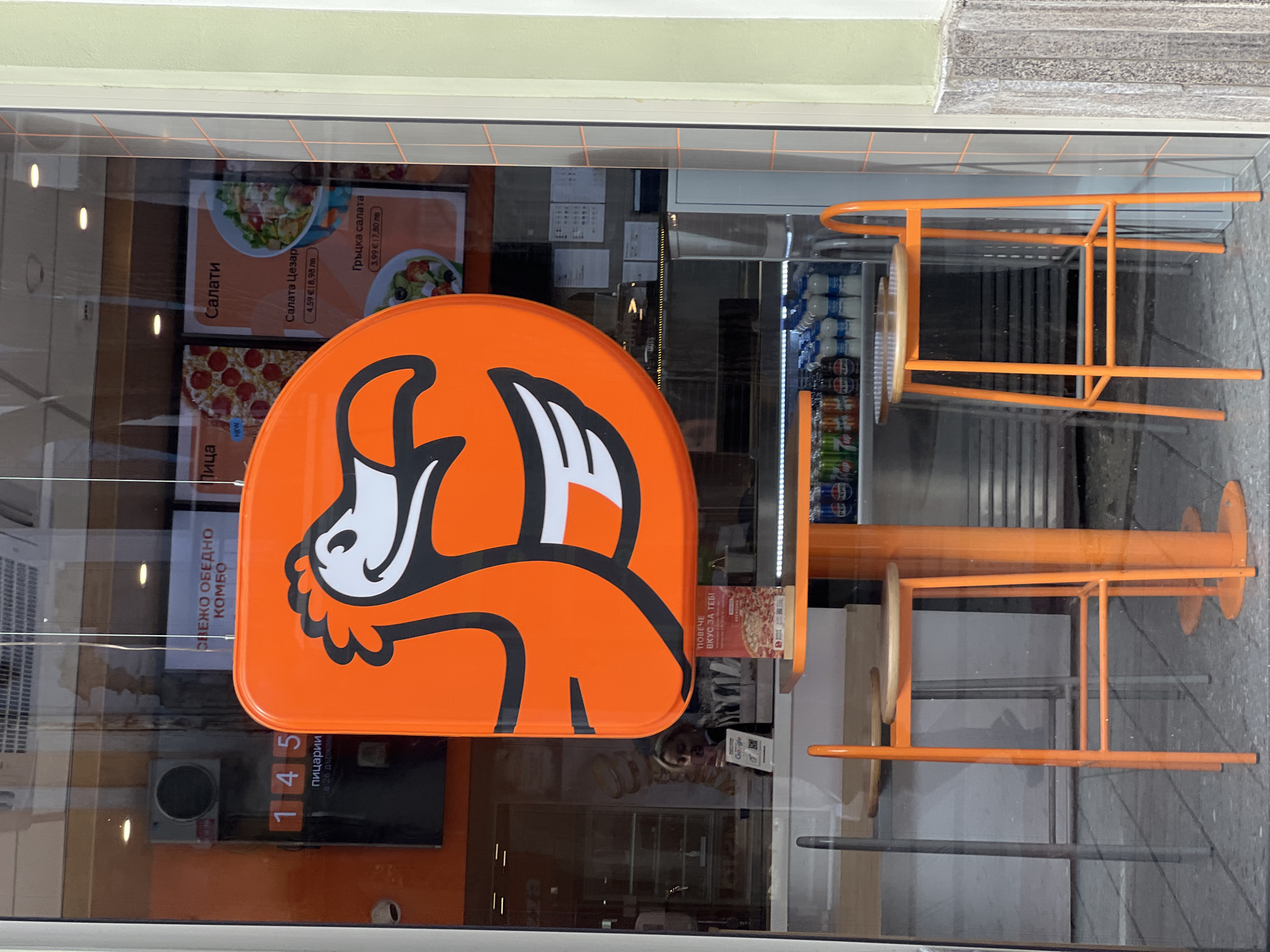 The image shows a storefront in Plovdiv, Bulgaria. An orange sign with a stylized white fox logo is prominently displayed on the glass window. Below the sign, a digital display reads "145 пицарии в 26 държа", likely indicating the number of pizza restaurants in 26 countries. Various posters advertising food options are visible in the window, including salads ("Салати") and pizzas ("Пица"), with prices listed in both Euro and Bulgarian Lev. An orange metal chair is stacked against the window on the right side of the image, reflecting the exterior street scene. The storefront appears to be part of a commercial building, with tiled surfaces above the window. A refrigerator filled with beverages is visible inside the store.