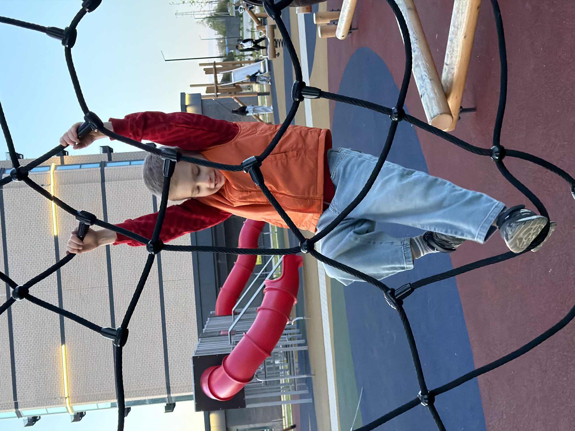 A young boy is actively climbing a black rope net structure at a modern playground in Yuzhny, Russia. The image is oriented sideways, depicting the boy holding onto the ropes with both hands and using his feet to support himself as if climbing upwards. He is wearing a red long-sleeved shirt, an orange puffer vest, light blue jeans, and dark sneakers with light soles. His eyes are slightly closed or squinted in concentration as he navigates the net.
The black rope net occupies a significant portion of the foreground, framing the boy. In the background, the playground features a vibrant, multi-colored rubberized surface in red, blue, and a beige-like tone. Various playground equipment is visible, including bright red tunnel slides with stairs and several wooden climbing structures. Other children can be seen playing on different elements of the playground in the middle ground. Beyond the play area, modern buildings with large windows and visible streetlights or poles extend towards the horizon, beneath a clear, bright sky, suggesting late afternoon or early evening. There is no visible text in the image.