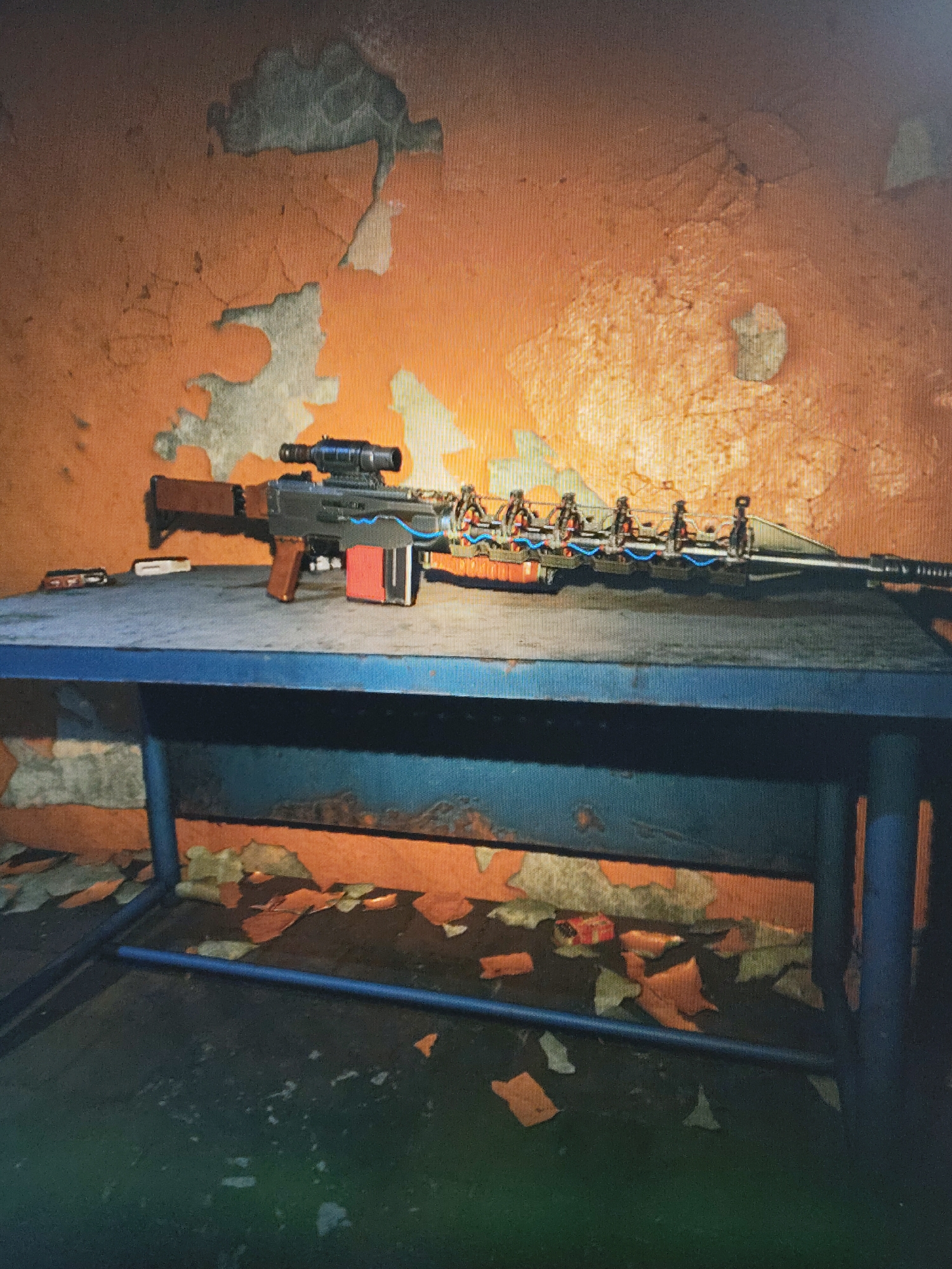The image displays a custom-built, modified rifle resting on a blue metal table. The rifle features a scope mounted on top, a wooden stock and pistol grip, and a prominent red magazine. A notable detail of the weapon is the intricate arrangement of blue and orange wiring connecting multiple small, cylindrical components along its fore-end, giving it a makeshift or experimental appearance.
The setting is an indoor space with a dilapidated aesthetic. The wall behind the table is painted orange, but large sections of the paint are peeling away, revealing a lighter, possibly concrete or plaster, layer underneath. This creates a distressed and aged texture. The floor beneath the table is dark and shows scattered debris, possibly more flakes of paint or other detritus, consistent with the worn state of the wall.
Small, indistinct items, possibly tools or other components, are visible on the table surface next to the rifle's stock. The lighting is somewhat dim, contributing to a gritty and unkempt atmosphere. The image itself appears to be a photograph of a digital screen, showing a slight pixel grid. No visible text is present in the image. The scene suggests a post-apocalyptic, derelict, or workshop environment, likely from a video game given the weapon's design and display context.