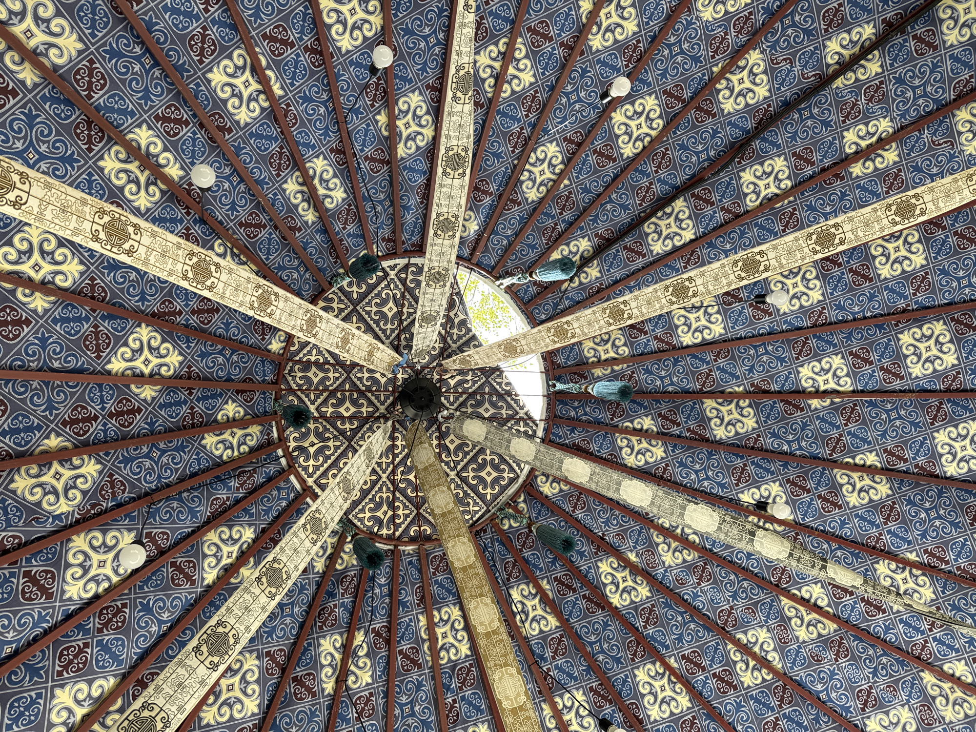 This is a low-angle shot of the ceiling of a yurt, likely in Leskhoz, Kazakhstan. The ceiling is covered in a patterned fabric with blue, yellow, and maroon motifs. Several wooden beams radiate from the center of the ceiling. Suspended from the beams are decorative fabric strips with intricate designs, some featuring tassels in shades of blue and green. There are also several exposed light bulbs providing illumination. The overall impression is one of traditional, possibly nomadic, architecture.