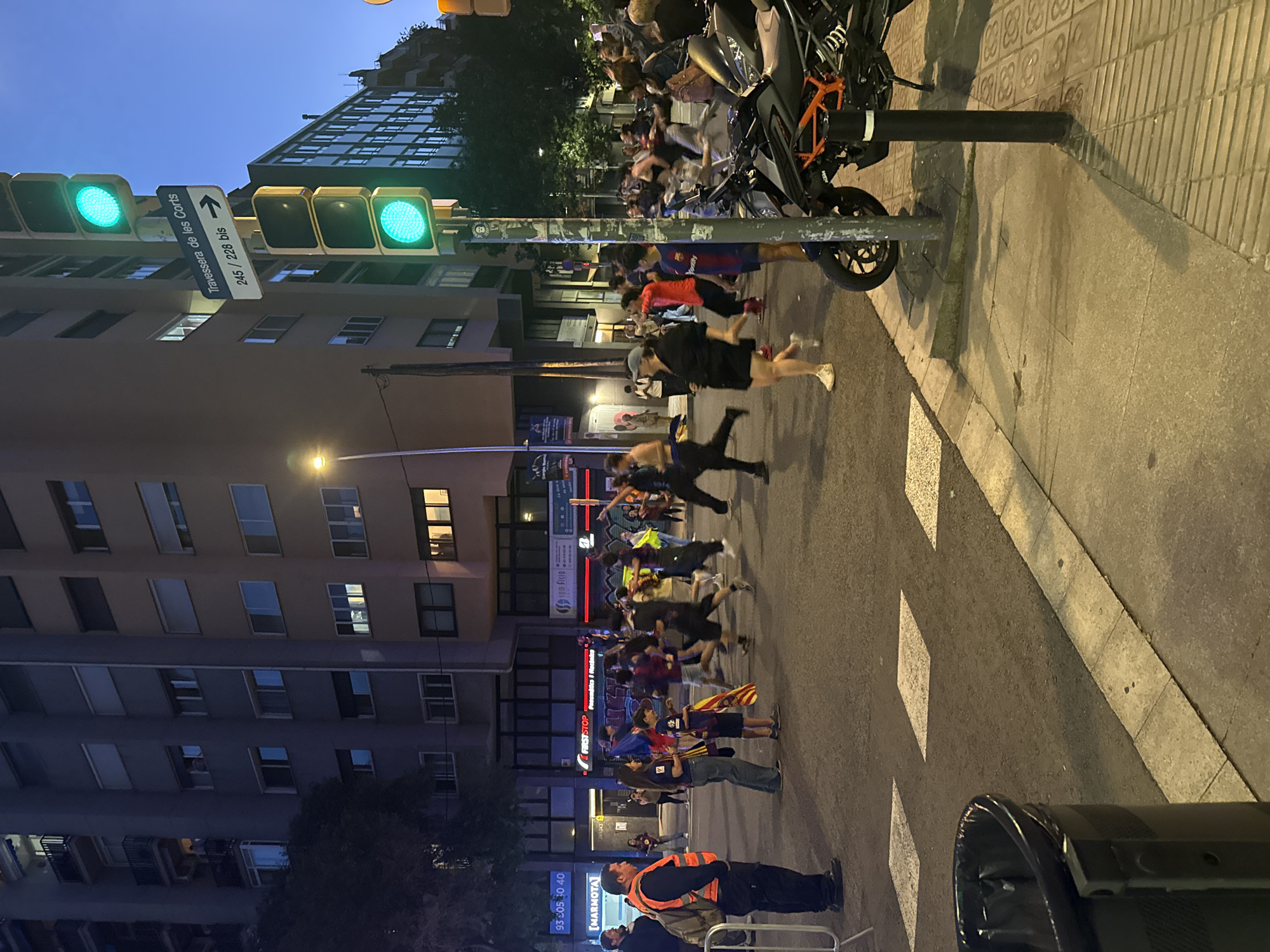 The image depicts a street scene in Barcelona, Spain, at dusk or night. A group of people, some wearing FC Barcelona jerseys and carrying Catalan flags, are running across the street. A security guard in an orange vest is standing near a barrier. A motorcycle is parked on the sidewalk. The traffic lights are green. A street sign indicates "Travessera de les Corts 245/228 bis." Business signage for FIRSTSTOP, MARMOTA (Pneumatics & Machaka), and Spotify can be seen on a building.