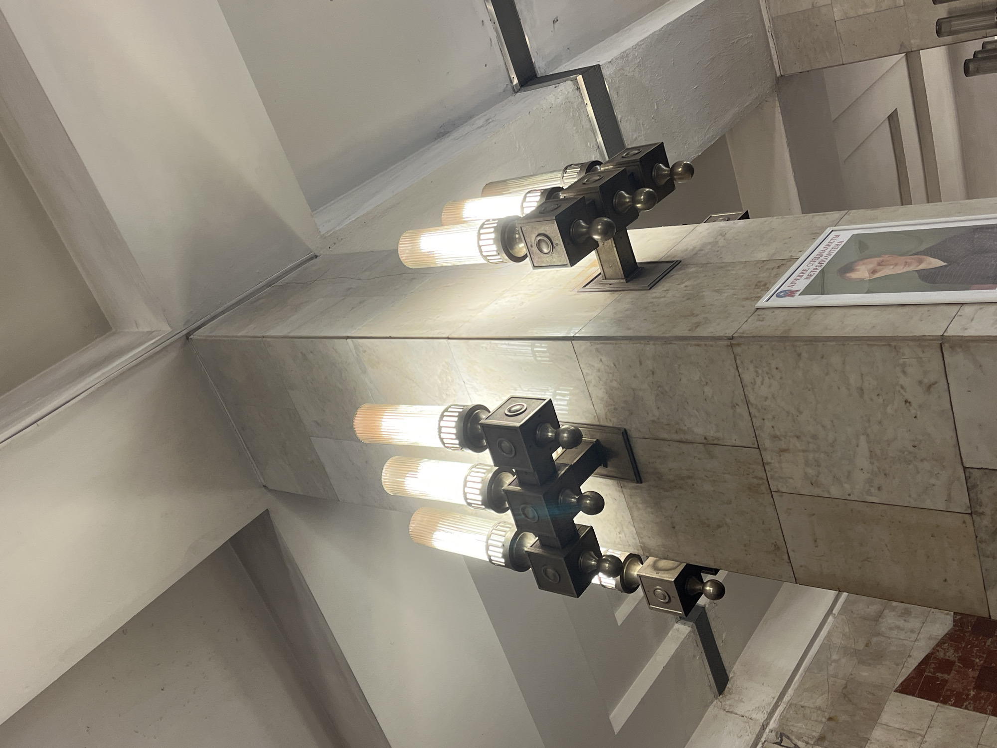 The media file shows an interior shot of a subway station, likely in Nizhny Novgorod, Russia, due to the Cyrillic text on a poster. The scene is illuminated by stylish, modern wall sconces, each featuring three cylindrical glass bulbs emitting a warm light. These sconces are mounted on a marble-tiled wall. Above the wall, a sloped ceiling forms geometric shapes with sharp angles and transitions into a plain white surface.
One of the sconces is positioned near a framed poster. The poster displays a portrait of a man and has Cyrillic text that reads "ЛУЧШИЕ СПЕЦИАЛИСТЫ МЕТРОПОЛИТЕНА" which translates to "BEST SPECIALISTS OF THE METROPOLITAN". This suggests an award or recognition for employees of the metro system.
The lighting creates a somewhat dramatic effect, highlighting the textures of the marble and the sleek design of the light fixtures. The overall impression is one of a well-maintained and modern public space. There are no people visible in the frame, and the time of day or weather conditions are not discernible from the interior setting. The focus is on the architectural details and the lighting design of the station.