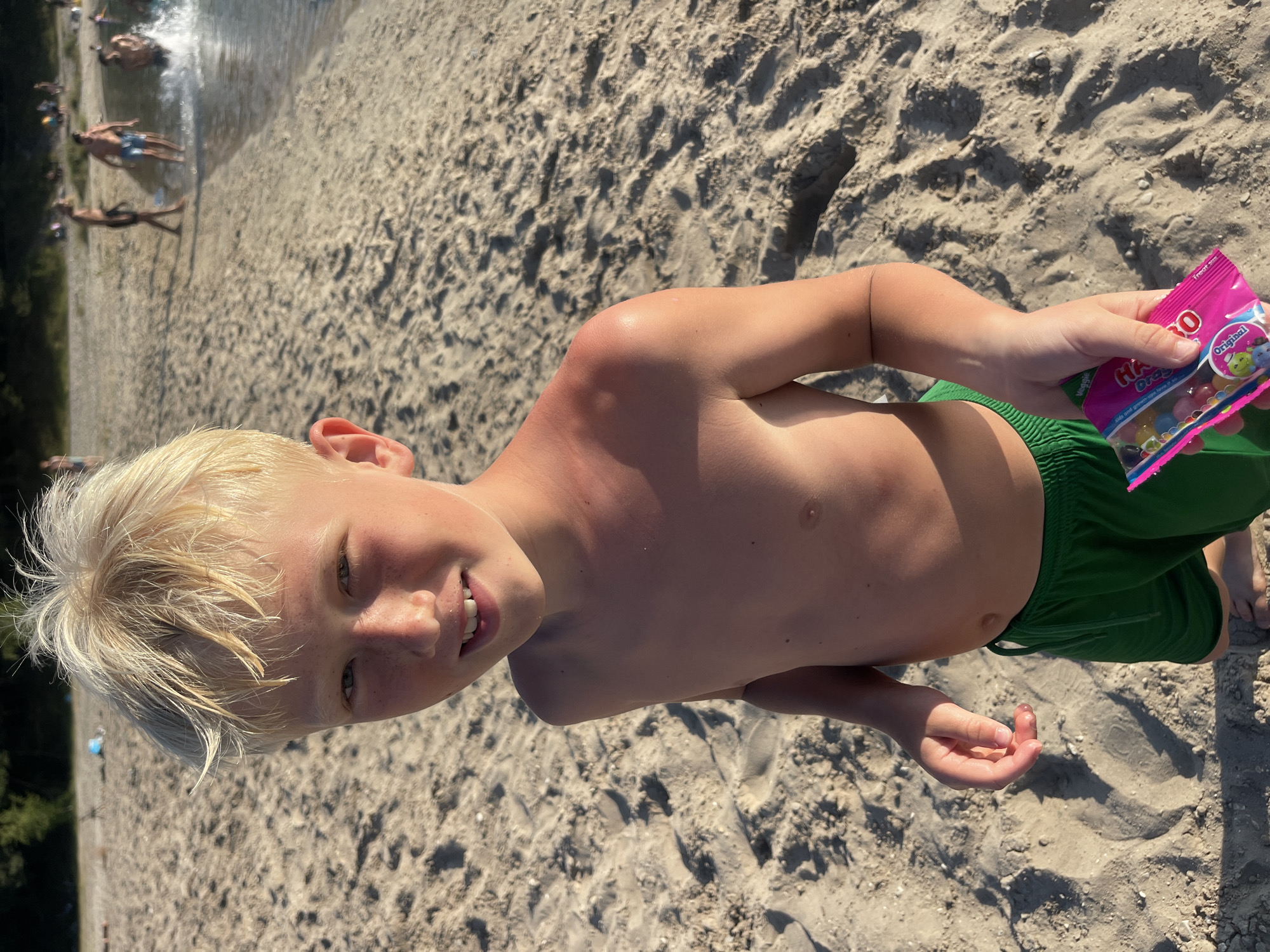 The image shows a young boy lying on a sandy beach, holding a bag of candy. He is shirtless and wearing green swim trunks. His face is turned towards the camera, and he is smiling. His skin appears to have some sunburn on his shoulder and upper arm. In the background, other people are visible near the water, some of whom appear to be swimming or wading. The scene is set on a sunny day, as indicated by the bright lighting and shadows on the sand. The sand is light brown and appears to be composed of fine grains. The water in the background is clear and reflects the sunlight. The bag of candy is pink and has text that includes "HARIBO" and "Original".
