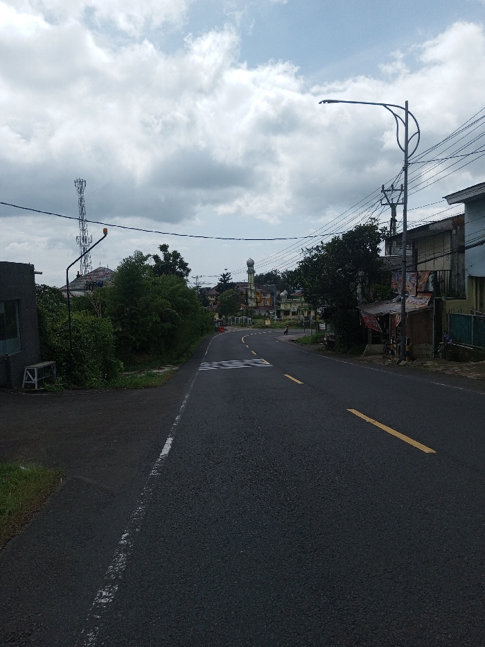This is an eye-level photograph of a paved road in Gubbisamamidi, India, under a cloudy sky. The asphalt road features a white solid line on its left edge and broken yellow lines in the center, indicating two-way traffic. A pedestrian crossing is marked with white stripes further down the road.
On the left side, a dark, low-rise concrete structure with a visible window or opening is present, next to which a white, wooden-framed bench rests. Beyond this, dense green foliage lines the road, obscuring the view of buildings. A tall, lattice-structured communications tower is visible in the upper-left, behind a multi-story building with a reddish roof. An overhead street light fixture with an orange indicator light is also positioned on the left.
The right side of the road features utility poles with multiple overhead electrical or communication lines. A modern street light on a curved pole extends over the roadway. Alongside the road, a series of buildings are visible, including one with a small, covered roadside stall displaying various items. In the far distance, a prominent mosque with a minaret and dome stands among other structures. A single, small, dark vehicle, likely a motorcycle or scooter, is faintly discernible on the road near the distant buildings. No individuals are clearly visible.