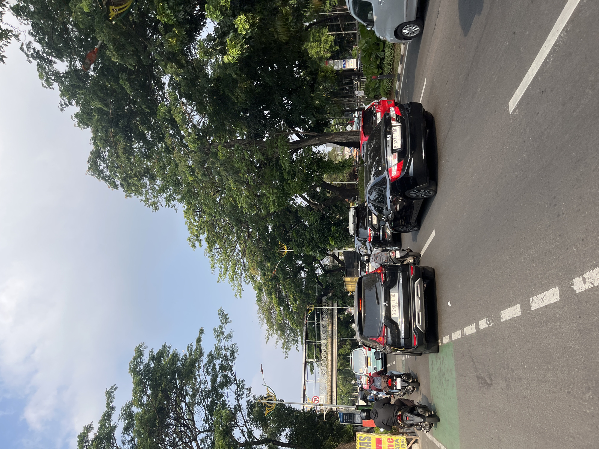 The image depicts a street scene in South Jakarta, Indonesia, characterized by moderate traffic on a clear day. The perspective is from a vehicle in the middle of the road, looking forward. On the right side of the road, a gray car is visible, partially obscuring a lane. Further ahead, a pedestrian overpass spans the road, supported by decorative pillars. Large trees with dense foliage line both sides of the street, casting shadows on the asphalt.
The road itself is marked with white dashed lines separating lanes. Several vehicles are present, including a black SUV in the foreground with license plate "B 2400 UOC." Behind it, a black sedan with license plate "B 1236 TAF" is also visible. To the left, two motorcyclists are riding in a designated green lane. One motorcyclist is wearing a black jacket and helmet, and the other is wearing a red backpack and helmet. A light blue van is stopped in traffic ahead of them.
On the far left, a yellow sign with red lettering is partially visible. The sky above is a clear blue with scattered white clouds. The overall impression is of a typical urban traffic environment during daylight hours.