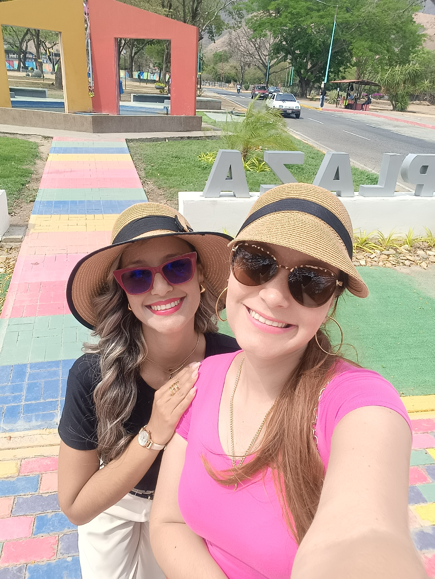 Two adult females are positioned in the foreground of an outdoor scene, posing for a self-portrait in a plaza in La Victoria, Venezuela. The woman on the right, closer to the viewer, wears a pink short-sleeved top, a straw bucket hat with a black band, and sunglasses with a tortoiseshell pattern. She is smiling, revealing teeth. The woman partially behind her on the left wears a black short-sleeved top, white pants, a wide-brimmed straw hat with a black band, and red-framed sunglasses. She is also smiling, showing teeth, and her right hand rests on the first woman's left shoulder. Her hand displays multiple rings and a watch.
Behind the women, large silver letters partially spell out "PLAZA". A paved walkway of multicolored rectangular blocks in pastel shades extends into the midground. To the left, two large, rectangular archway structures, one yellow and one red, stand on a concrete base. Beyond these elements, a paved road runs horizontally across the midground, with a white car and a partial view of a brown pickup truck. Several light poles with blue accents are visible along the road. On the far side of the road, a brown-roofed, open-sided structure has several people present. The background features green trees and distant hills or mountains under a clear sky. The environment suggests a sunny day.