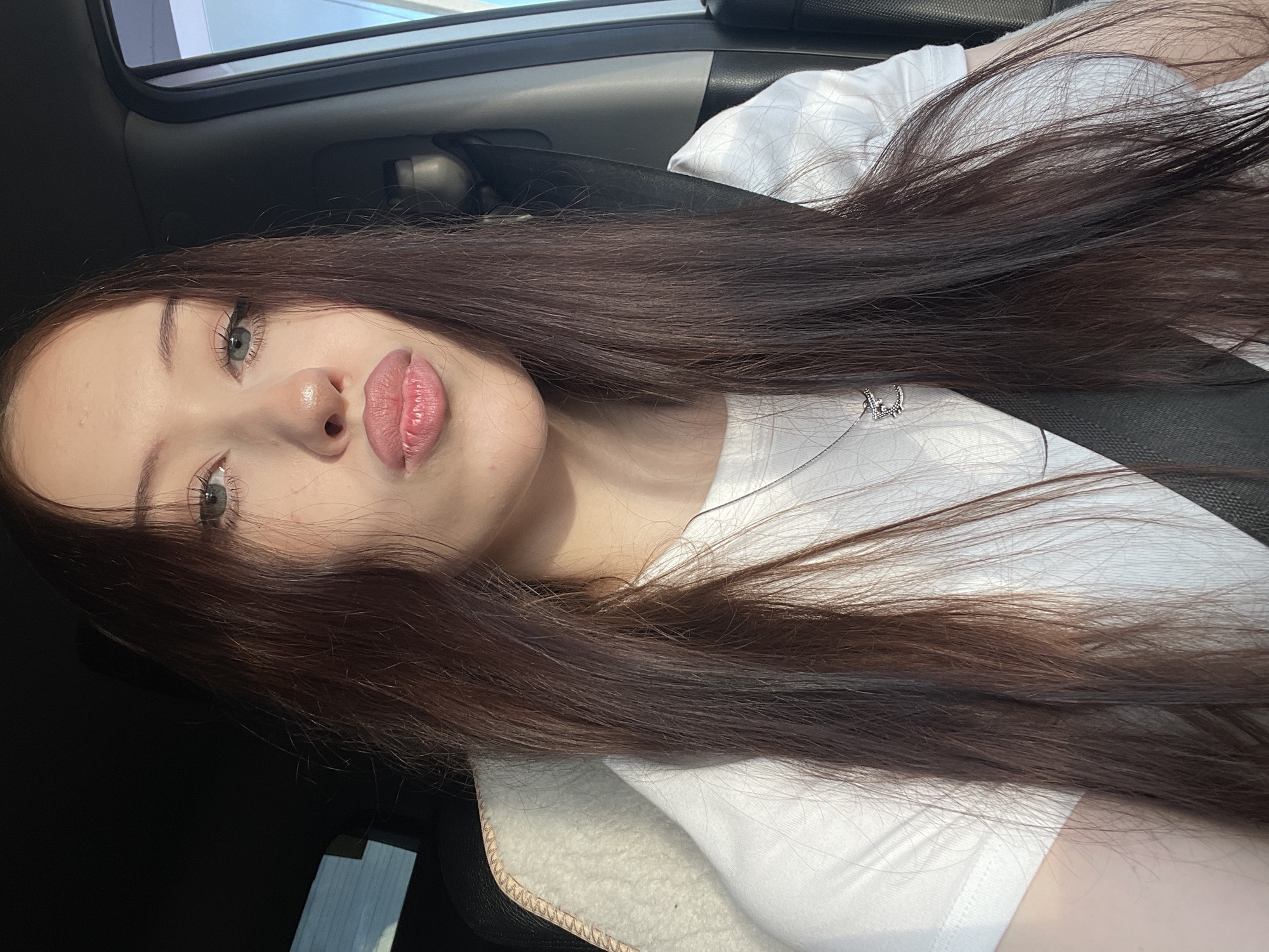 The media file is a portrait photograph taken in a car, with the subject's head and shoulders prominently featured. The subject is a young person with long, dark brown hair that extends across the frame. They have fair skin, light-colored eyes, and full lips with pink lipstick. A delicate silver necklace with a small pendant is visible around their neck. The subject is wearing a white t-shirt.
The setting appears to be inside a vehicle, suggested by the car's interior elements like the seatbelt and the textured material of the seat. The lighting suggests it is daytime, possibly with sunlight filtering in. The overall mood of the image is relaxed and casual, with the subject looking directly at the camera. There is no visible text in the image. The provided location context is Гавриловка, Russia.