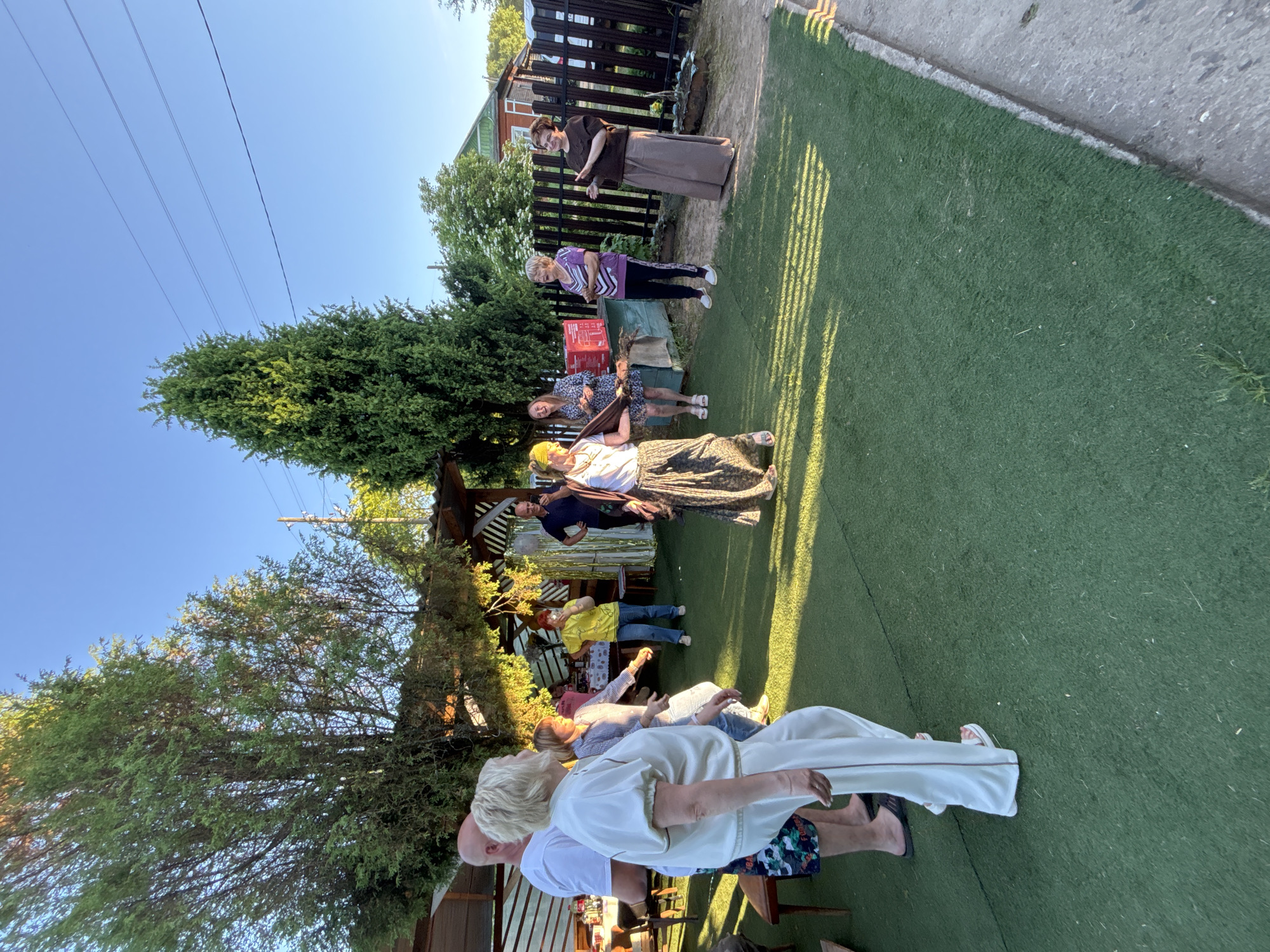 This image depicts an outdoor social gathering of approximately ten adults in a backyard setting in Rozhdestveno, Russia, during the late afternoon or early evening, evidenced by the long shadows cast across the ground. The majority of the visible ground is covered with artificial green turf, with a concrete edge on the far right.
In the mid-foreground, a woman with short blonde hair wears a white long-sleeved top and white wide-leg trousers with white sandals, facing right and appearing to step or dance. To her immediate left, a bald man wears a white t-shirt and patterned shorts. Behind them, a woman in a light blue and white checkered shirt and blue jeans also moves to the right. Further right, near the center, a woman with a yellow headscarf, white top, and long patterned skirt actively dances with arms slightly outstretched, facing left.
Other attendees are observed in various states of interaction and observation. To the left, a woman in a yellow t-shirt and blue jeans watches. To the right of the dancing woman, a woman with short blonde hair wears a purple and black striped top and black trousers, smiling. On the far right, a woman in a dark brown long-sleeved top and long light brown skirt gestures with her hands, facing the group. In the background, under a wooden pergola, a man in a dark blue polo shirt stands observing.
The environment includes mature green trees, notably tall conifers on the left and center. A wooden pergola or gazebo is visible on the left, possibly with a table holding drinks. On the right, a black metal fence borders the property, behind which a house with a green roof and brown walls is partially visible, along with a light-colored car and additional greenery. A small wooden structure holding a red box is situated near the fence. The sky is clear and light blue, traversed by multiple power lines in the upper right quadrant.