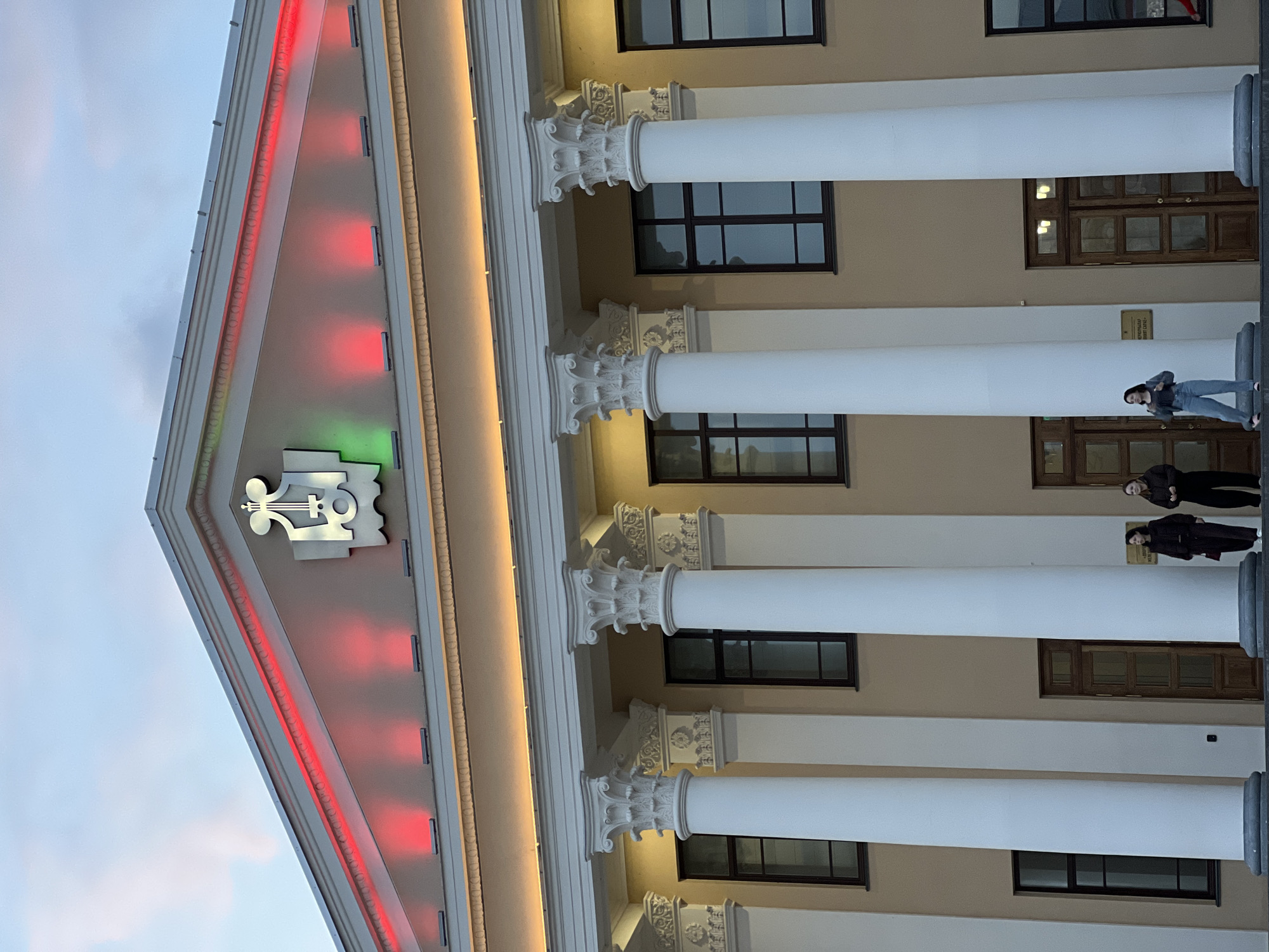The image depicts a building facade in Kazan, Russia, specifically the Saydashev State Grand Concert Hall. The building features classical architectural elements including a pediment with red and green lighting along the sides and a musical emblem. Four prominent white columns stand in front of the building. Behind the columns are windows and wooden doors. Two people are standing on the platform in front of the entrance. One person is wearing jeans and a jacket. The other is wearing darker clothing. The sky is visible in the upper left corner.