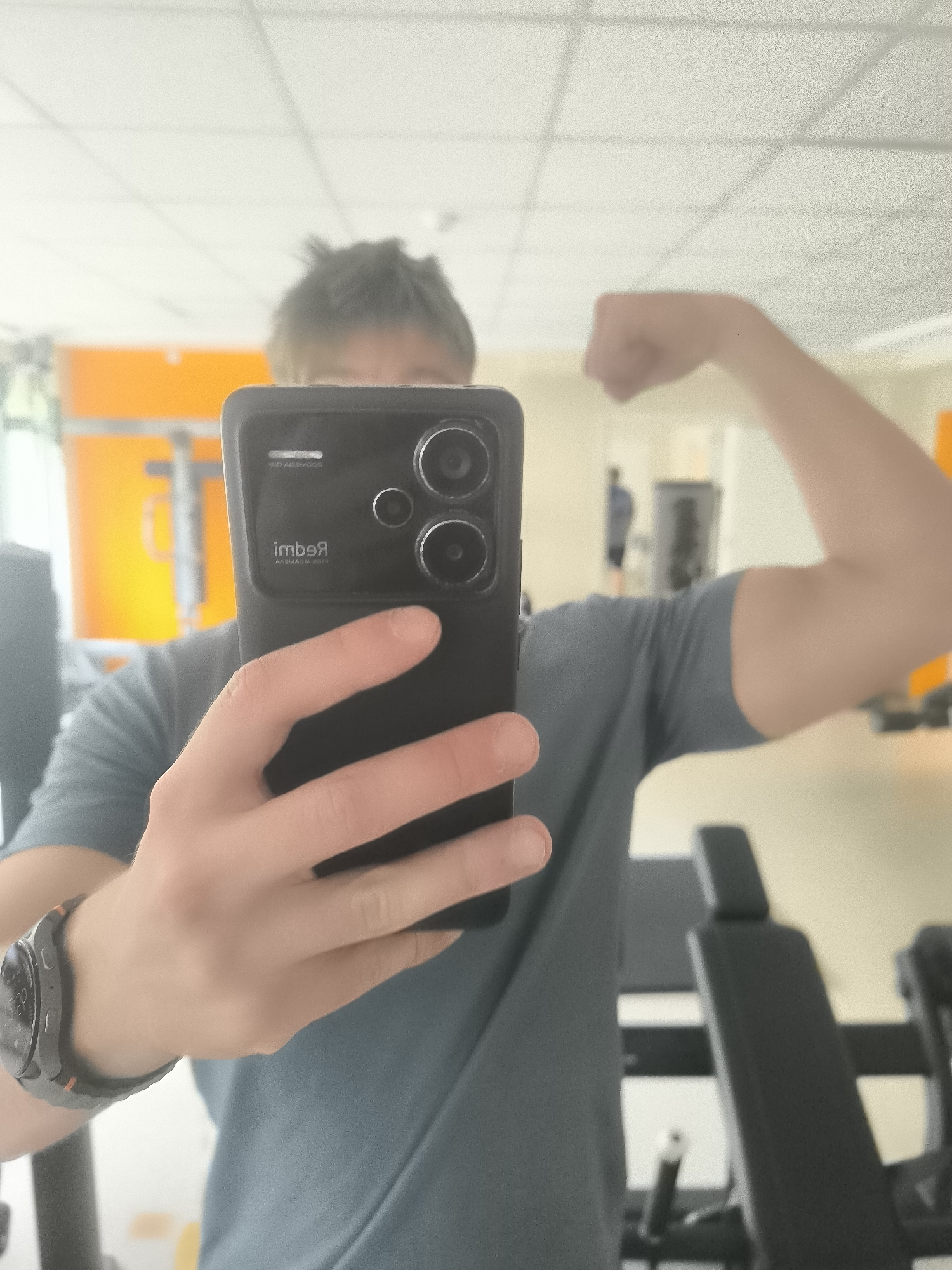 A selfie is taken in a gym, showing a person with short brown hair wearing a grey t-shirt and a black smartwatch on their left wrist. The person is holding a black smartphone in their left hand, obscuring their face. Their right arm is flexed, showing a slightly muscular bicep. The background shows gym equipment, including weight benches, and an orange wall with a rack of weights. A person can be seen walking in the background. The gym has a neutral-colored floor and ceiling with visible lighting panels. The phone camera shows the word "Redmi" in reverse. The location is likely in Sofia, Bulgaria, given the user's request.
