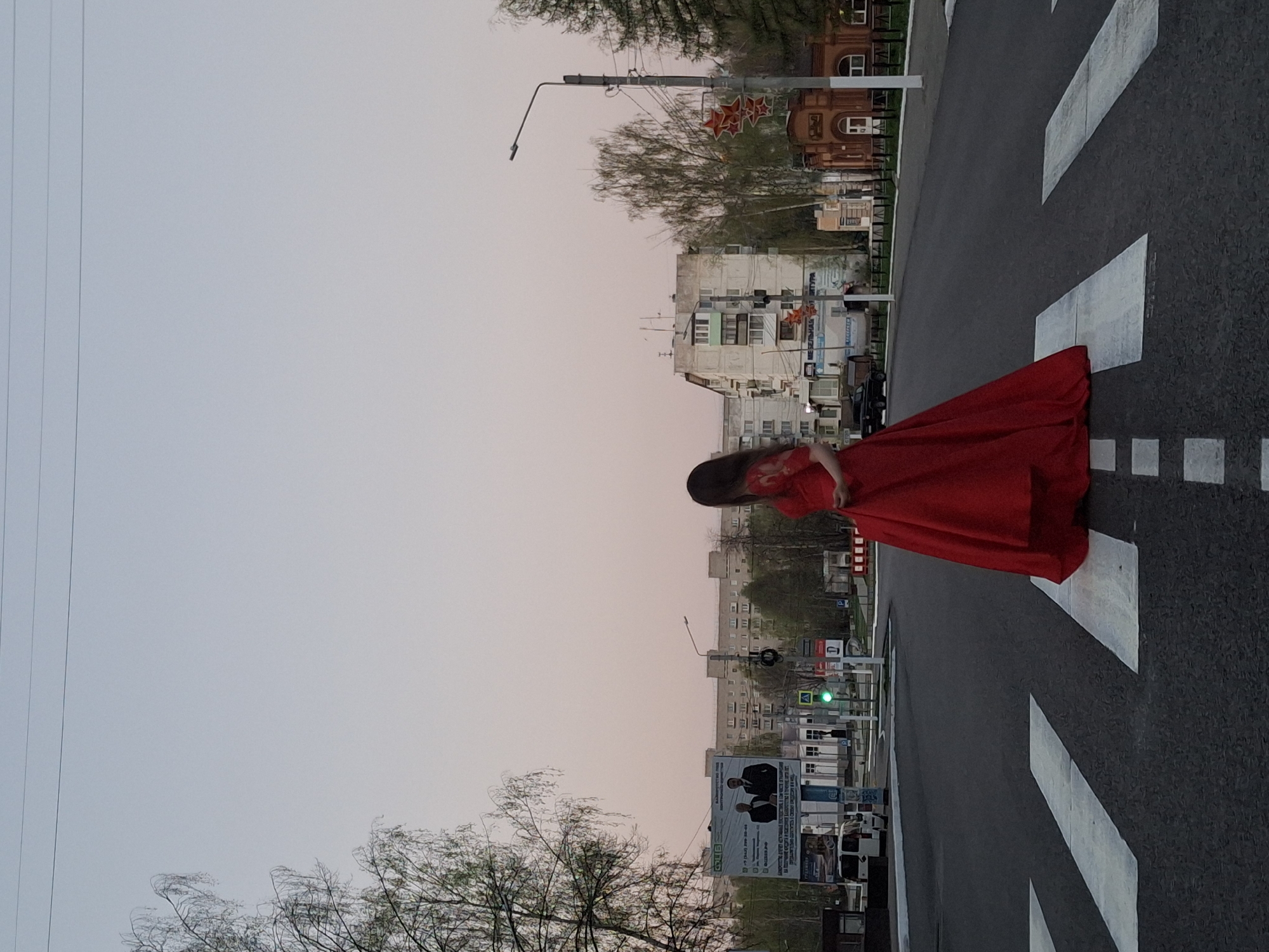 A woman in a red dress is standing on a crosswalk in a city, possibly Chaikovsky, Russia, based on background details like building style and signage. The crosswalk is marked with white stripes. The sky is a light pink hue, suggesting dawn or dusk. Buildings line the street on either side, with trees interspersed. Power lines are visible in the sky. A street lamp is present, with what appears to be a red star emblem on it, a symbol common in Russia. A large billboard with writing and images is visible. A traffic light shows a green signal.