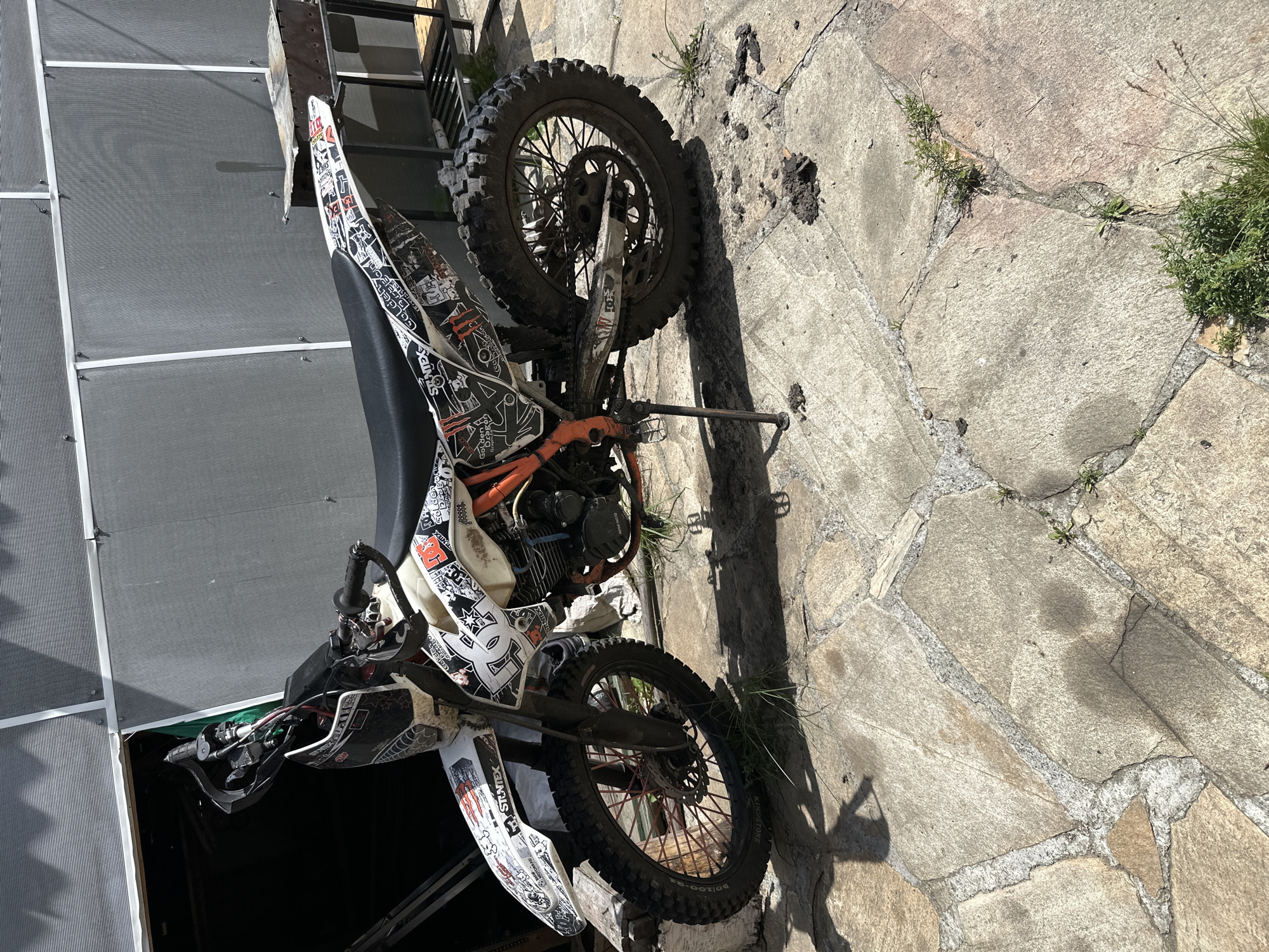 This media file depicts a dirt bike parked outdoors on a paved stone surface. The dirt bike is predominantly white with numerous black and red stickers and graphics covering its body. The stickers include various brand logos and designs, such as "DC Shoes," "Monster Energy," and what appears to be a spiderweb design.
The bike has knobby tires suitable for off-road use, with the rear wheel and suspension clearly visible. The engine and some orange-colored components of the bike are also visible. A kickstand is deployed, supporting the bike.
The setting appears to be an outdoor area with a stone patio or walkway. Some weeds and grass are growing in the cracks between the stones, suggesting it might be a slightly unkempt or natural outdoor space. In the background to the left, there is a section of a grey, textured surface, possibly a screen or fence, and part of a metal structure, like stairs or a railing.
The lighting suggests it is daytime, with strong sunlight casting shadows, particularly from the bike onto the stone ground. The shadows indicate the sun is positioned to the side. There is no visible activity taking place; the bike is stationary. The overall impression is one of a parked dirt bike in an outdoor, possibly residential or recreational, environment.