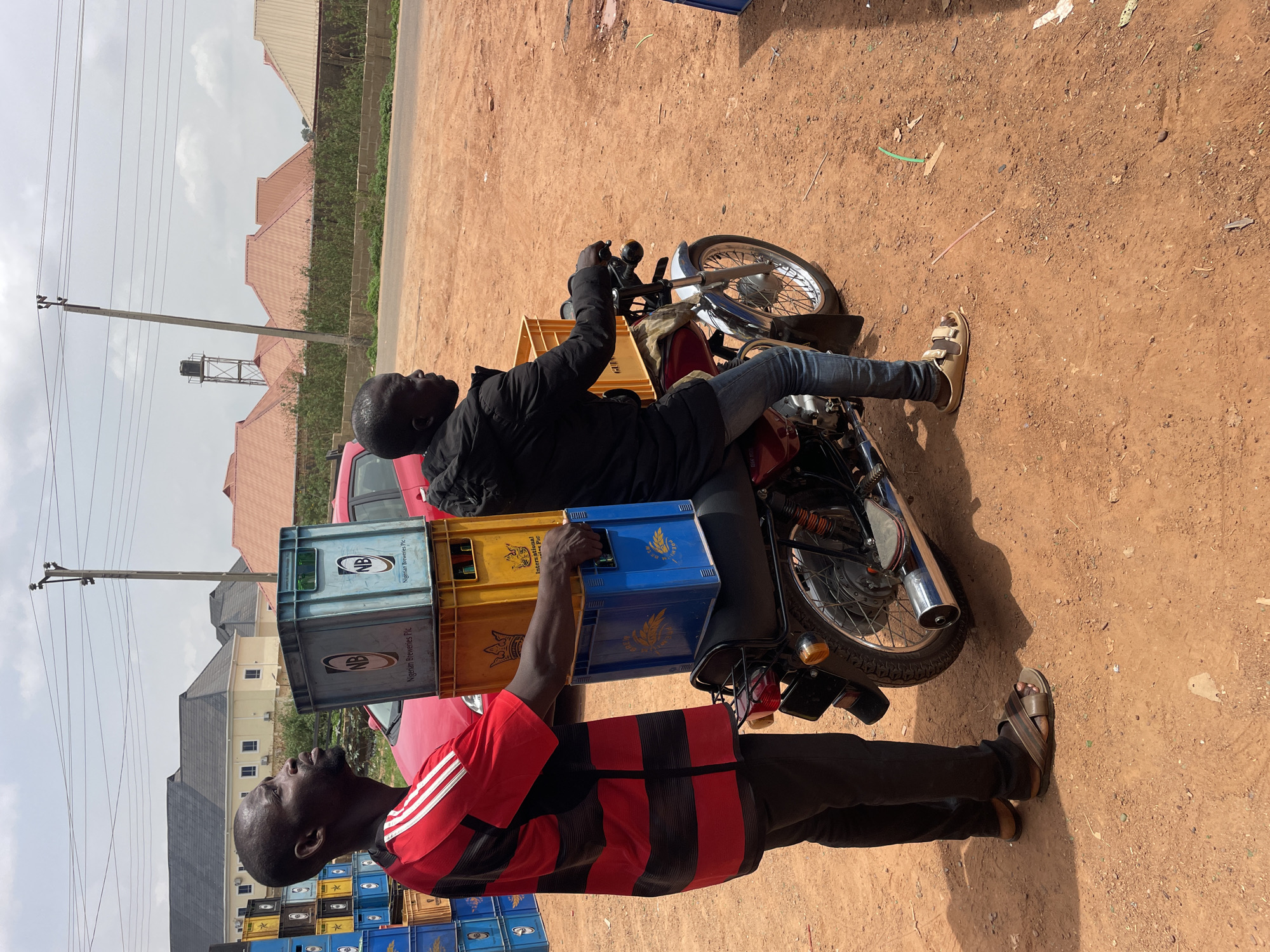 A man wearing a red and black striped shirt is standing beside a motorcycle loaded with stacked crates in Gura Top, Nigeria. A second man is seated on the motorcycle. The crates appear to contain bottled beverages. The background consists of a dirt road, buildings, and a cloudy sky.