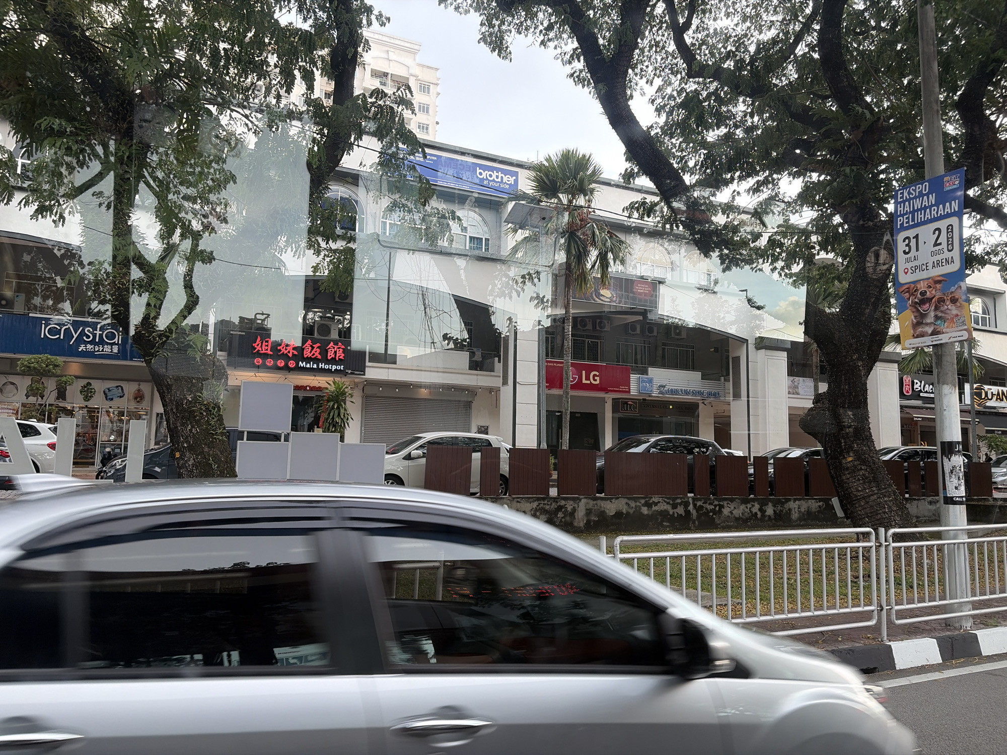 The image shows a street scene in an urban environment, likely in Malaysia due to the presence of Malaysian language on a sign. A silver car is in the foreground, partially obscuring the view of a building behind it. The building appears to be a commercial establishment with glass storefronts. Signs for businesses such as "brother", "Mala Hotpot", and "LG" are visible. Trees with dense foliage are along the street, casting shadows. A lamppost with a blue and white banner advertising an event is also present. The overall atmosphere suggests a daytime scene on a cloudy day.