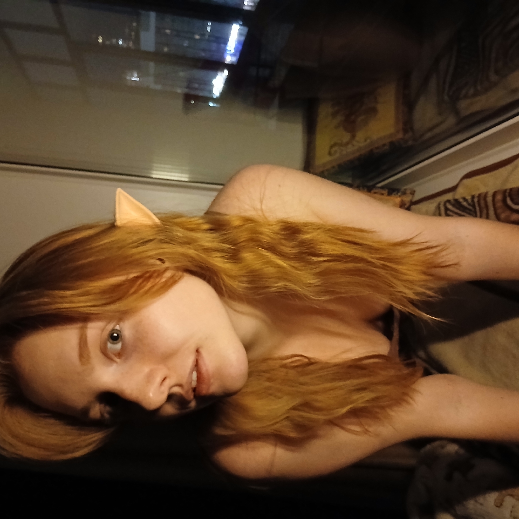 This image, set in Yekaterinburg, Russia, features a young woman indoors at night. She has long, wavy reddish-blonde hair and light-colored eyes, and is looking towards the viewer with a slight smile. A pair of light-toned, pointed elf ears are worn as an accessory, protruding from her hair. Her shoulders and upper chest are visible, suggesting she is partially dressed or wearing a low-cut top.
The woman is lying down, possibly on a bed or couch, with a patterned fabric in shades of brown and cream visible to her right. A large window behind her reflects blurred city lights, indicating an urban setting at night. Through the reflection, a hint of an ornate, possibly framed artwork or tapestry in gold and dark tones can be seen on an interior wall. The lighting is generally dim, contributing to the nocturnal ambiance. There is no visible text in the image.