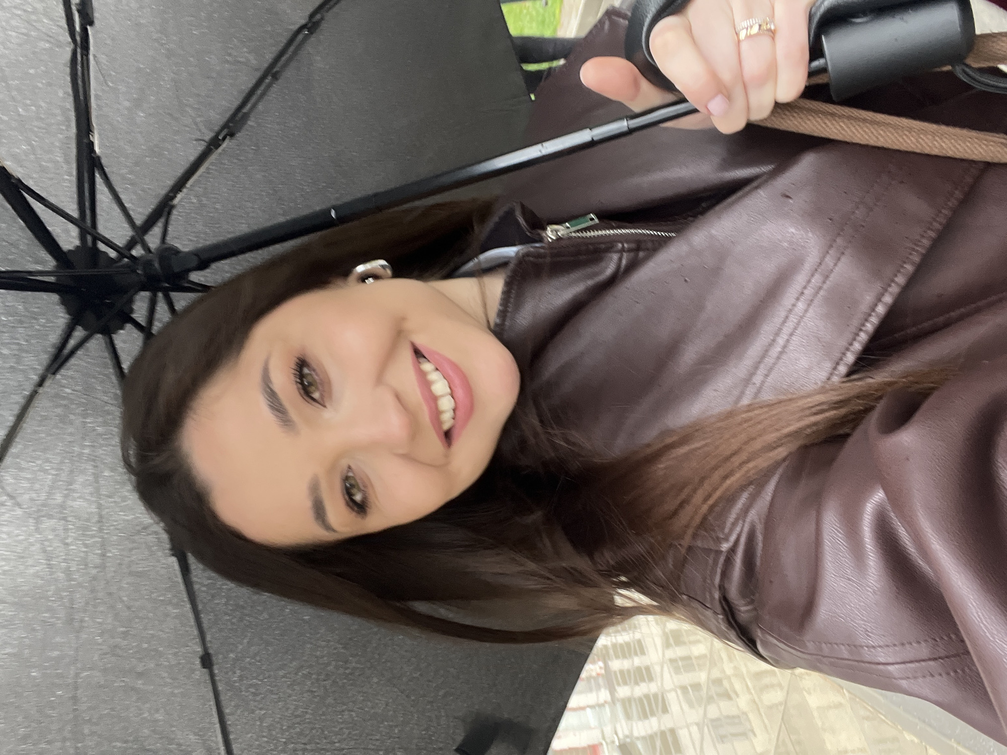 A woman with long, dark brown hair is posing indoors under an open black umbrella. She is wearing a brown leather jacket. She has fair skin and is smiling, showing her teeth. She has visible rings on her left hand. The location is presumably in Khimki, Russia.