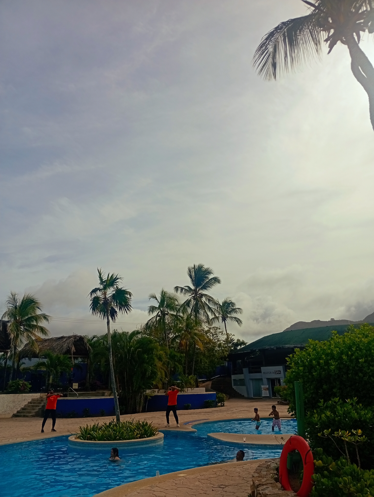 The photograph depicts a resort swimming pool at Playa El Agua, Venezuela. The pool is a multi-tiered, lagoon-style design with a bright blue interior. Several people are in and around the pool: some swimming, others wading, and two individuals in matching orange shirts appear to be leading activities on the pool deck. The pool area is surrounded by palm trees and tropical foliage, with a building visible in the background bearing the sign "Hesperia SPA." Beyond the building, a low, green mountain range can be seen under a cloudy sky. A red life preserver is positioned near the pool's edge. The ground surrounding the pool is paved with light-colored stone tiles.