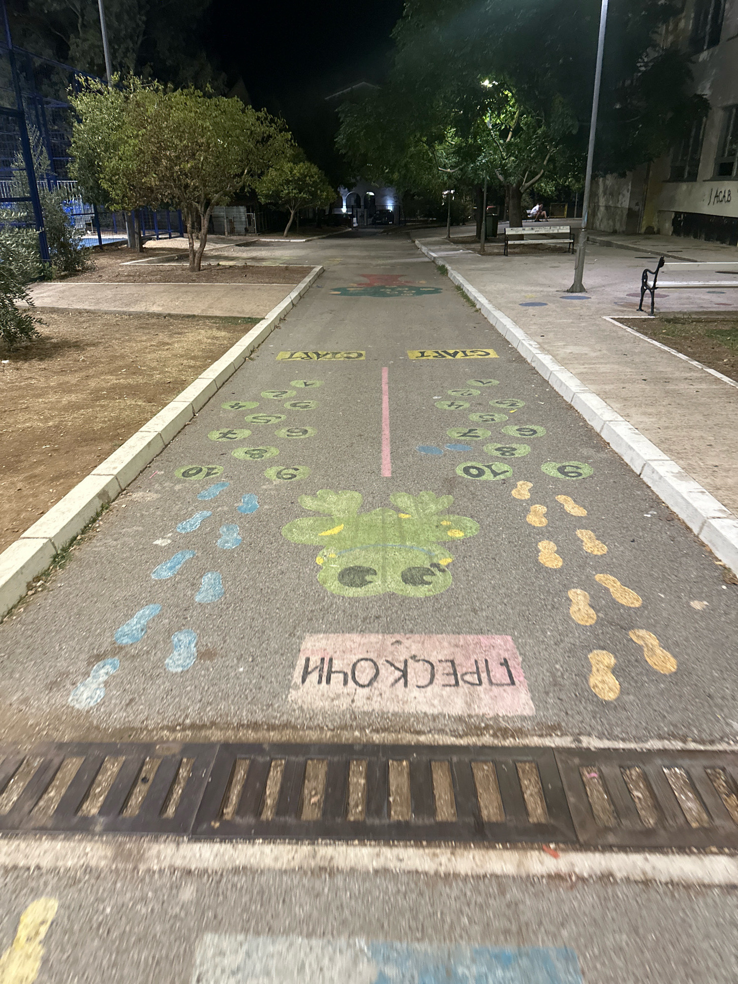 This image depicts a hopscotch court drawn with chalk on a paved walkway at night. The court features numbered circles from 1 to 10 in green, along with painted footprints in blue and yellow on either side. A stylized green frog is painted in the center. In the background, trees line the walkway, and streetlights illuminate the scene. A metal grate is visible in the foreground. Graffiti, including "J ACAB", is present on a building in the far background. The presence of a hopscotch court and the general urban park setting suggests this is likely in a public recreational area.