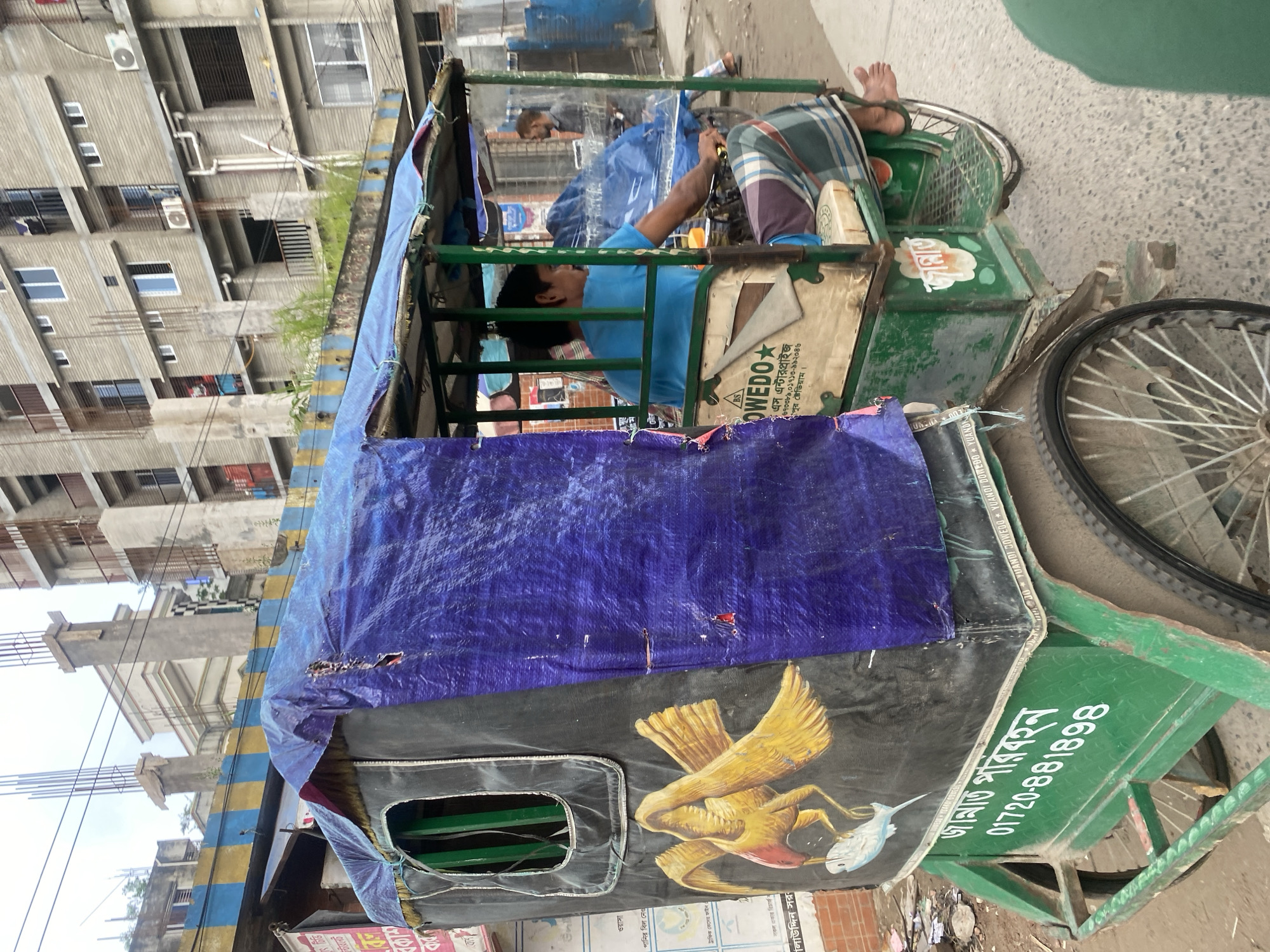 A bright green and black rickshaw, decorated with a painting of a yellow bird holding a white fish and a purple tarp covering its roof, is parked on a street in Dhaka, Bangladesh. A young man in a blue t-shirt and a striped lungi is sitting in the driver's seat, looking ahead. In the background, there are multi-story buildings under construction, with exposed concrete and rebar. The street is paved with concrete and there are some trash piles on the side. A man in a grey shirt is standing in the distance, facing away from the camera. The overall scene depicts a typical street view in a developing urban environment.