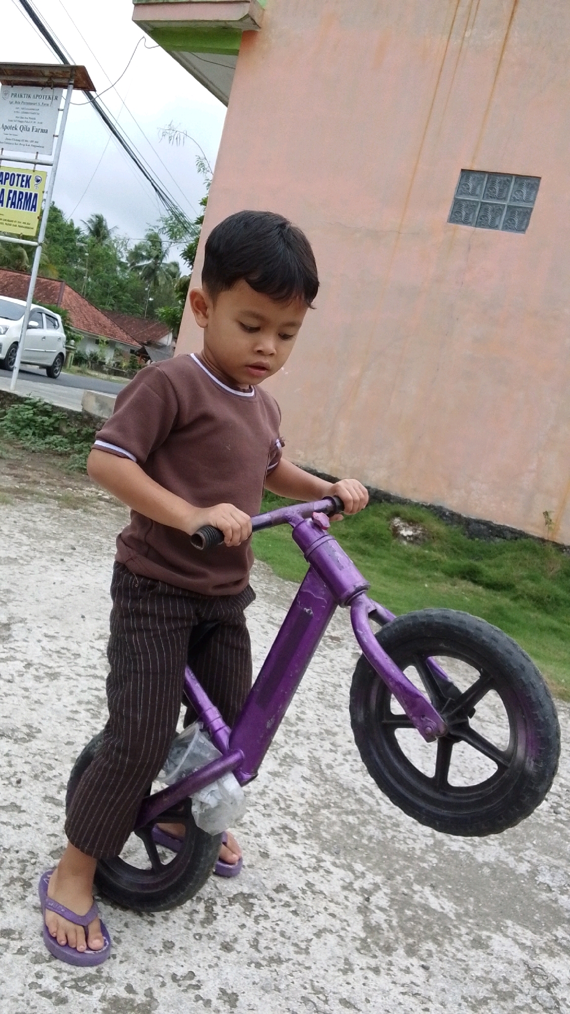 A young boy, appearing to be of Southeast Asian descent, is captured in a full-body shot, standing outdoors on a concrete surface. He is dressed in a brown t-shirt with white trim on the collar and sleeves, paired with dark brown striped pants and purple flip-flops. The boy is holding onto the handlebars of a purple balance bicycle. The bicycle is small, suitable for a child, with black tires and a silver reflector attached to the front. The boy is looking down at the bicycle.
The background reveals a building with a light pinkish-orange painted wall, featuring a small window made of frosted glass blocks. To the left of the building, a signpost stands, displaying text that appears to be in Indonesian, with the words "PRAKTIK APOTEKER" and "APOTEK QILA FARMA" visible. A white car is parked on a road behind the signpost, and beyond that, residential buildings with red tile roofs and lush green trees are visible, suggesting a rural or suburban setting. The sky above is overcast.
The image's context points to Cintaratu, Indonesia, as indicated by the signage. The overall scene depicts a candid moment of a child with his bicycle in a seemingly ordinary day.