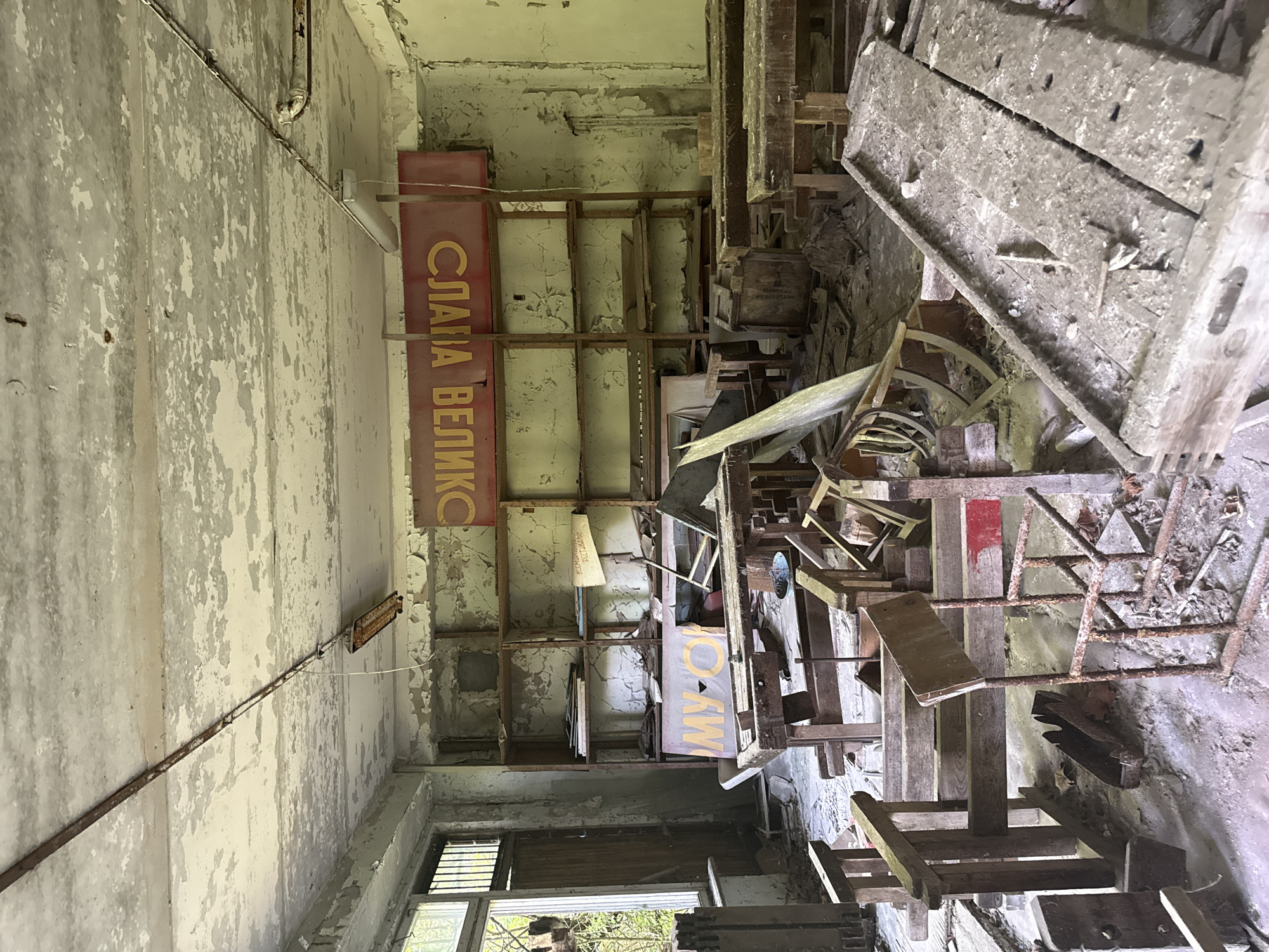 The image depicts the interior of a dilapidated room, likely an abandoned workshop or storage area. The scene is cluttered with various discarded objects, primarily made of wood and metal, suggesting a past industrial or craft-related use. These items are stacked haphazardly, creating a sense of disarray and neglect.
Dominating the left side of the frame is a red sign with Cyrillic lettering in yellow. The visible text reads "СЛАВА ВЕЛИК" which translates to "Glory to the Great". Another sign with similar yellow lettering on a lighter background is partially visible further back in the room, reading "МУО". The walls and ceiling are peeling, showing layers of aged paint and dampness, indicative of long-term disuse. A rusted metal pipe runs along the left wall, connected to a fluorescent light fixture.
The lighting suggests it is daytime, with natural light entering from a window visible on the lower right. The overall atmosphere is one of decay and abandonment, evoking a sense of history and perhaps melancholy. The space appears empty of people. The arrangement of objects and the signage hint at a Soviet-era context, possibly a factory, school, or community center that has fallen into disrepair.