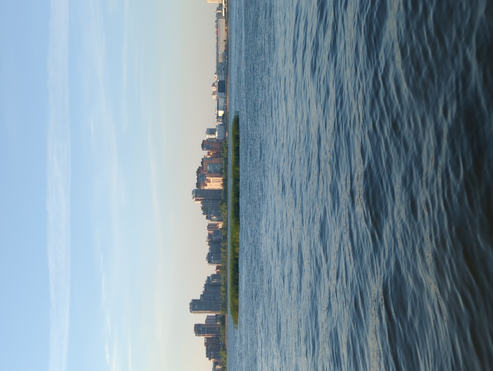 The image depicts a panoramic view of the city of Kazan, Russia, on a clear day with a blue sky. In the foreground, the surface of a body of water, likely the Kazanka River or the Kuybyshev Reservoir, is visible with gentle ripples. In the middle ground, a small island covered in reeds or tall grass stands out in the water. Beyond the island, the cityscape of Kazan unfolds, featuring a cluster of modern high-rise buildings with varying architectural styles and colors. Some buildings are made of glass and concrete, while others have facades of brick or stone. The buildings are densely packed, indicating a developed urban area. The background shows a clear horizon with a pale sky, suggesting it is either early morning or late afternoon. No people or vehicles are explicitly visible in the image. The overall scene conveys a sense of urban development integrated with natural elements.