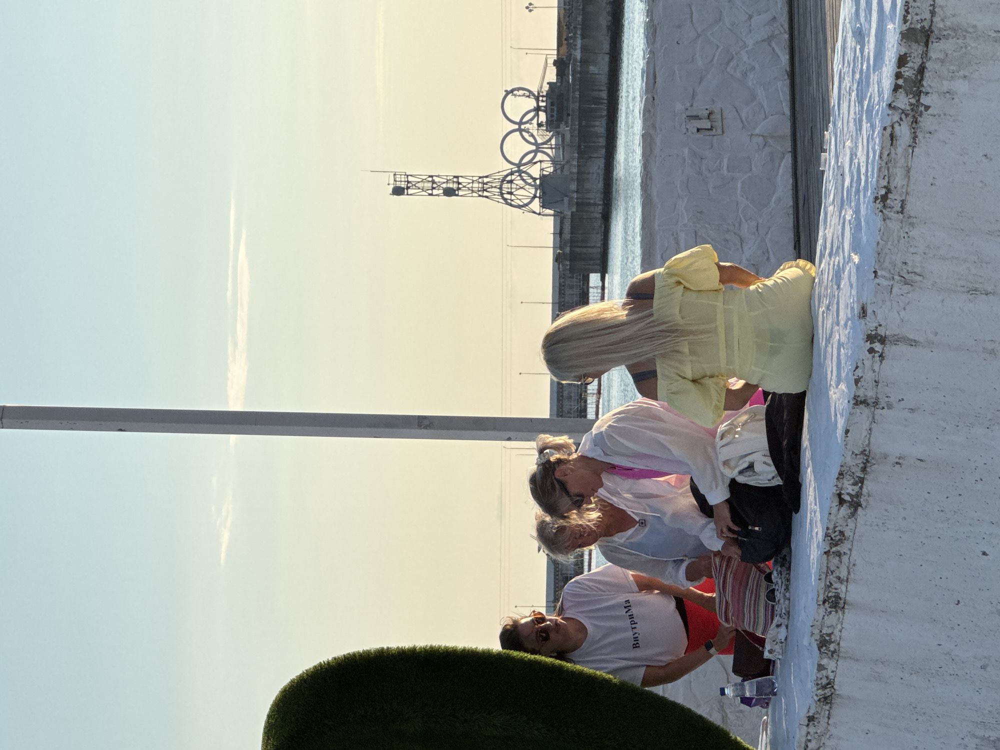 The image shows a group of people gathered near a body of water, with a large Olympic rings structure visible in the background, hinting at a location in Sochi, Russia. The scene appears to be set outdoors during what seems to be late afternoon or early evening, given the warm, soft lighting and long shadows cast. The sky is a pale blue with a touch of yellow near the horizon, suggesting clear or slightly hazy weather.
The subjects are a mix of individuals, some appearing to be adults, engaged in various activities. One person in the foreground is wearing a white t-shirt with Cyrillic text that reads "ВнутриМа" which translates to "Inside Mama". This person is also wearing sunglasses and appears to be looking downwards. Other individuals are dressed in casual attire, including a woman in a light yellow dress. Some are seen interacting with each other, possibly resting or engaged in conversation.
Objects present include a water bottle on the ground and a striped bag. The background features a textured white wall and the Olympic rings structure, which is a prominent landmark. The overall atmosphere seems relaxed and informal. The presence of the Olympic rings strongly suggests the location is Sochi, a city known for hosting the 2014 Winter Olympics.