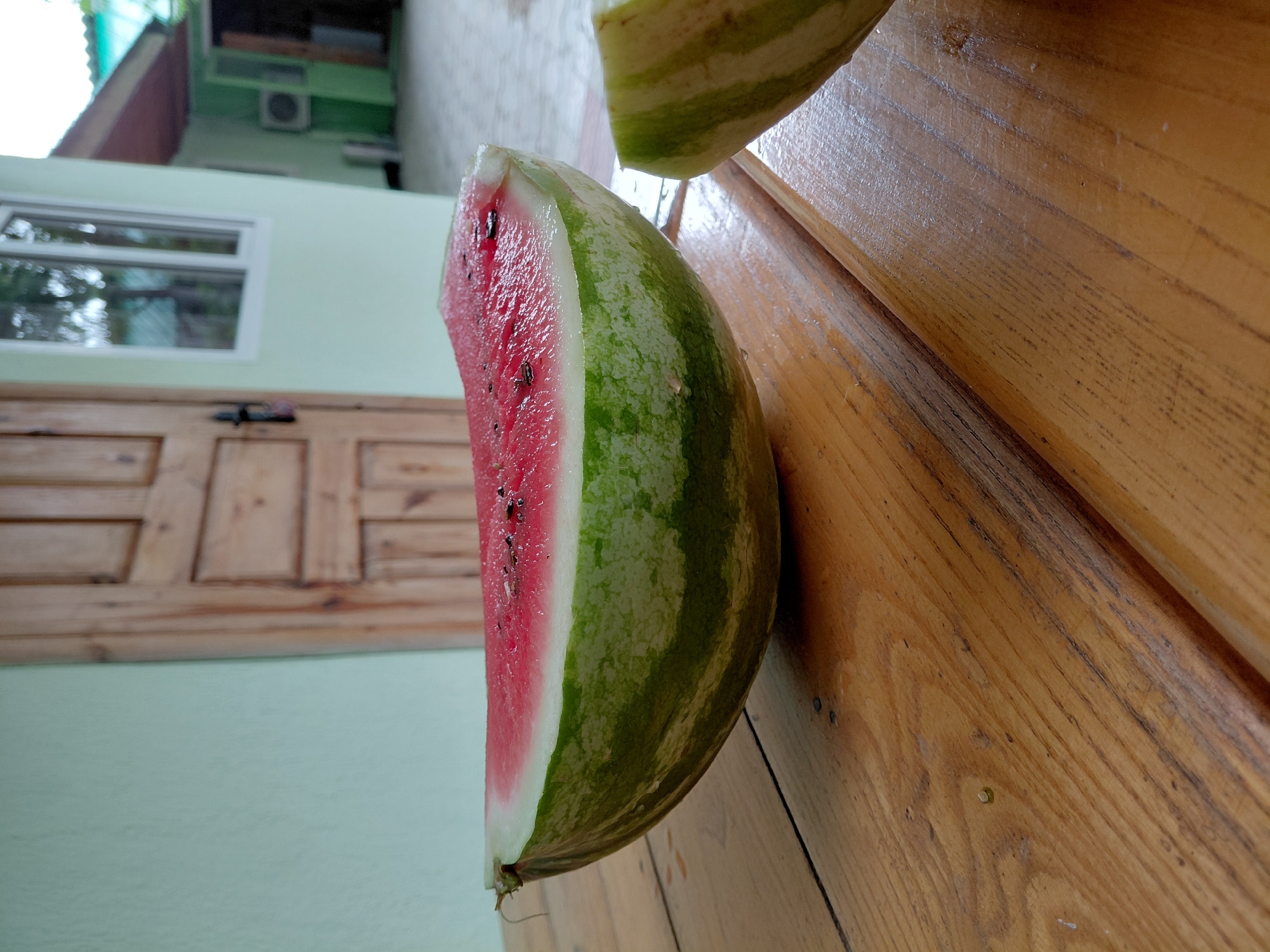 A sliced watermelon rests on a wooden surface, its vibrant pink flesh and green rind sharply contrasting with the warm tones of the wood. Another portion of the watermelon, also sliced, is visible in the upper part of the frame. The background depicts an outdoor scene with a light green wall, a wooden door with a window, and part of a building. The lighting suggests it is daytime. The context provided indicates the location is Akshi, Kazakhstan. There is no visible text in the image.