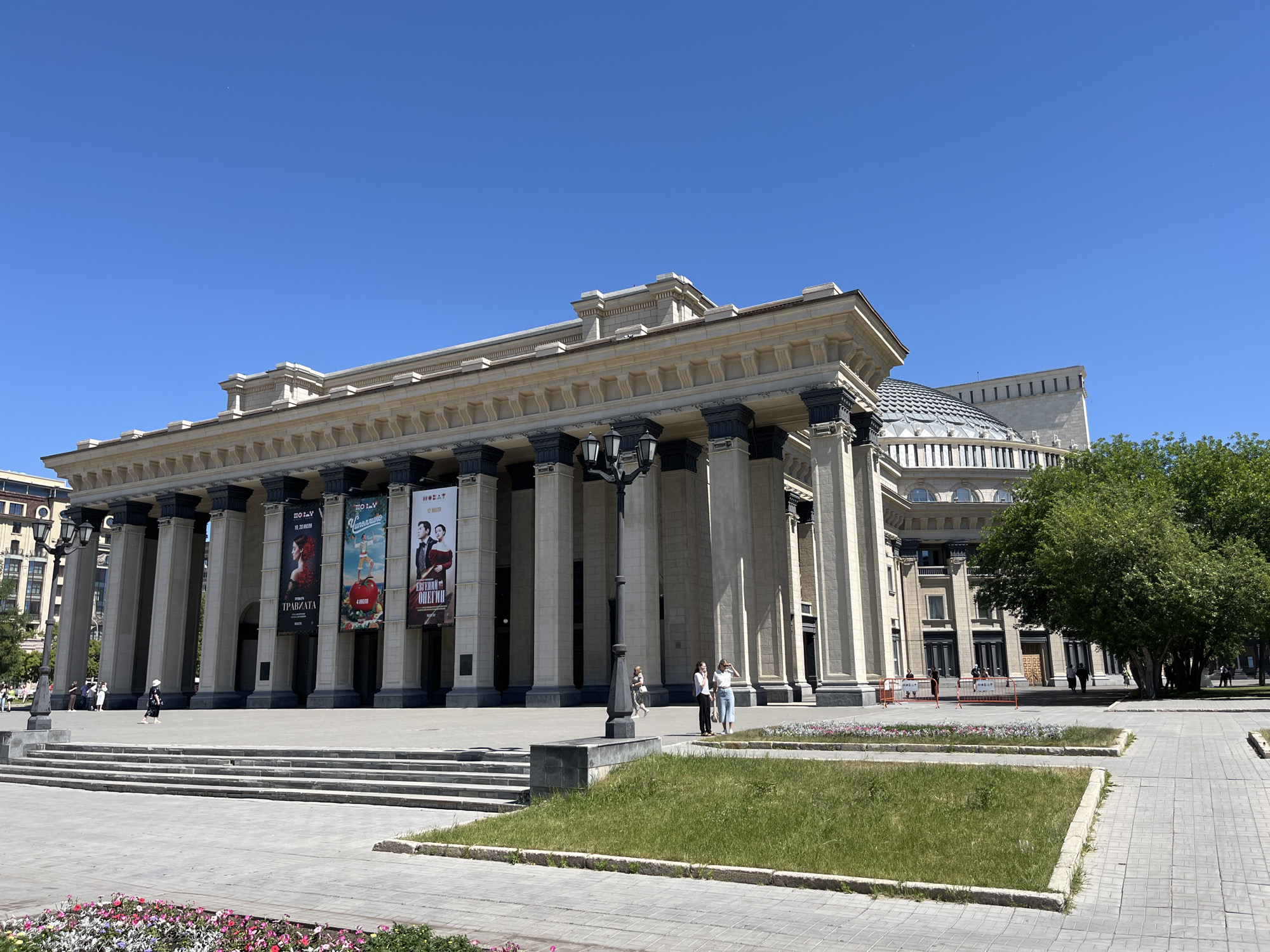 A wide, eye-level, outdoor, daytime shot shows the neoclassical facade of a large, light-colored public building. The building features a grand, colonnaded entrance with numerous tall, dark-colored pillars. Hanging banners with images and text are displayed on the facade. To the right of the entrance is a large, domed section of the building with a glass roof. A lush green tree is situated to the far right of the building. In front of the building, there is a wide, paved plaza with several tiered concrete steps leading up to the entrance. Several people are visible walking in the plaza and on the steps. The sky is a clear, bright blue. Flowerbeds with colorful flowers are visible in the foreground.