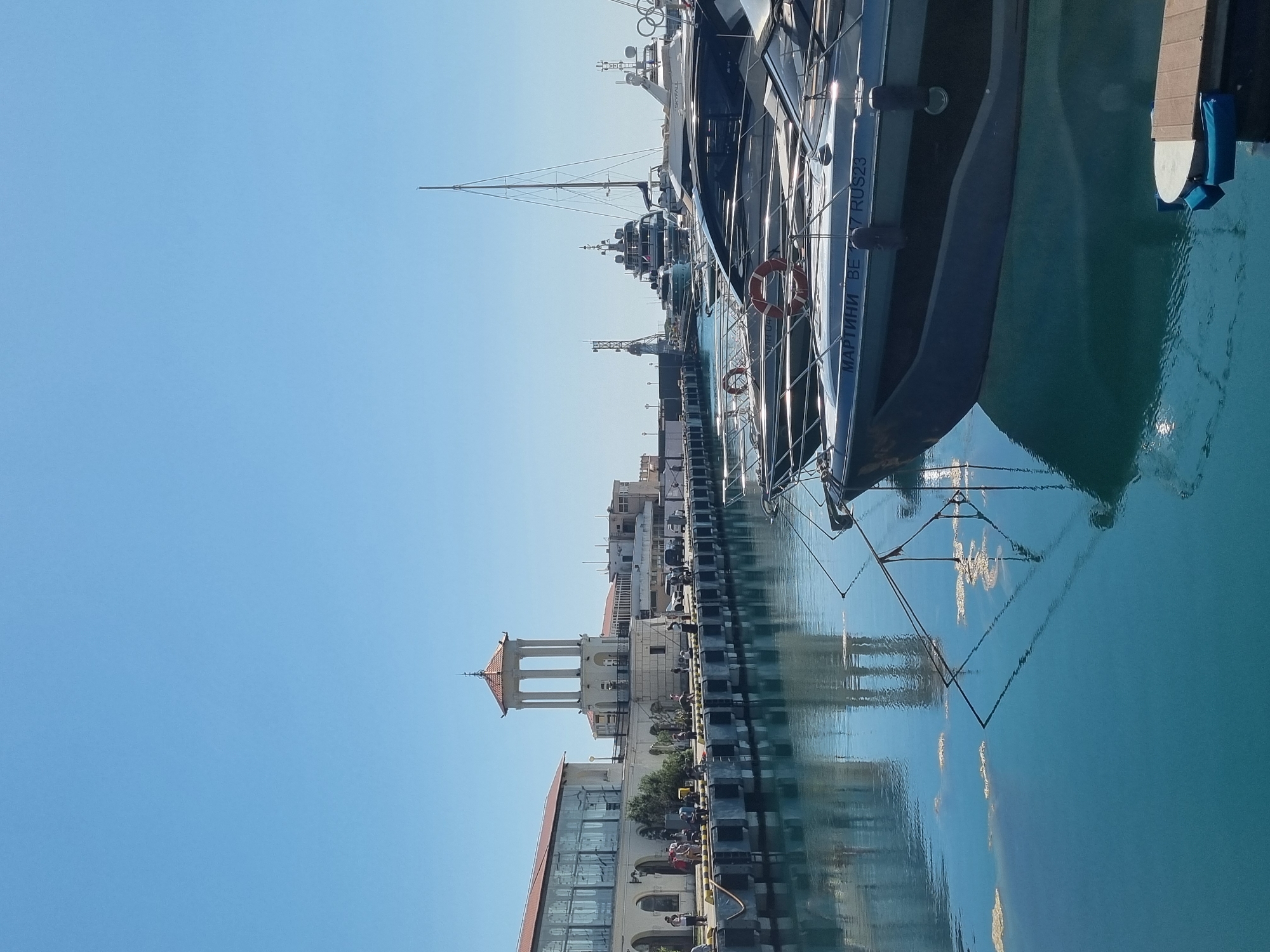 The image depicts a waterfront scene, likely a harbor or marina, in Sochi, Russia. The foreground features several boats docked along a concrete pier. One boat is prominently positioned on the right side of the frame, with its name partially visible in Cyrillic script. The boats are reflected in the water, indicating calm conditions.
In the background, a building with a tower stands out, with a red tile roof. Several other buildings, mostly of light color, line the waterfront. The sky is clear and blue.
