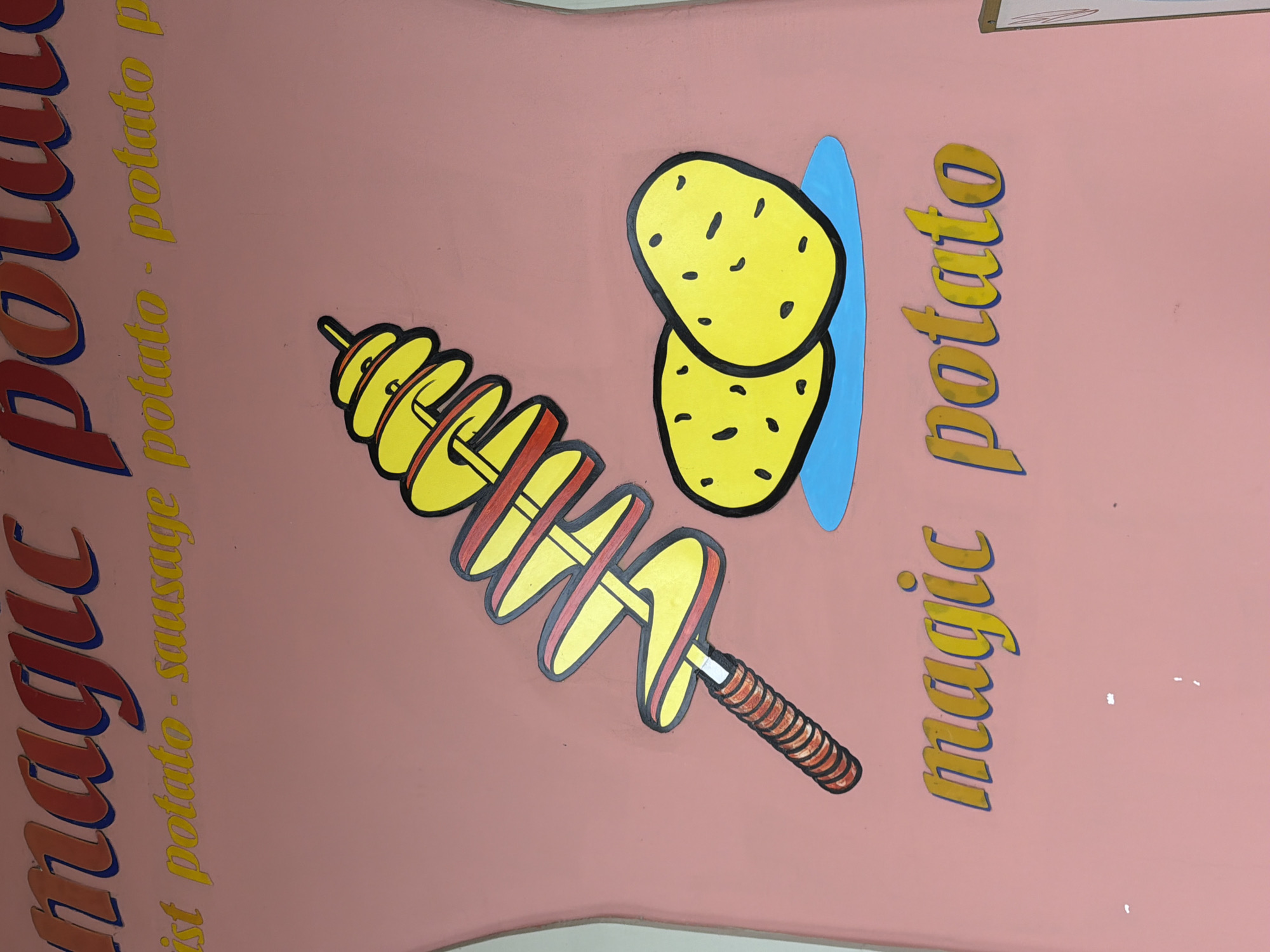 The image is a close-up of a pink wall decorated with text and cartoon illustrations of potatoes. At the top, in large red and blue lettering, is the word "magic". Below this, in yellow cursive script, are the words "ist potato - sausage potato - potato p". To the right of the text, a cartoon depicts a spiral-cut potato on a stick and two whole potatoes drawn in a simple yellow style with black dots for eyes. Below these illustrations, in yellow lettering outlined in blue, are the words "magic potato". The wall appears to be part of an outdoor advertisement or mural, possibly for a food stall in Đà Nẵng, Vietnam, specializing in potato dishes. The overall style is playful and vibrant.