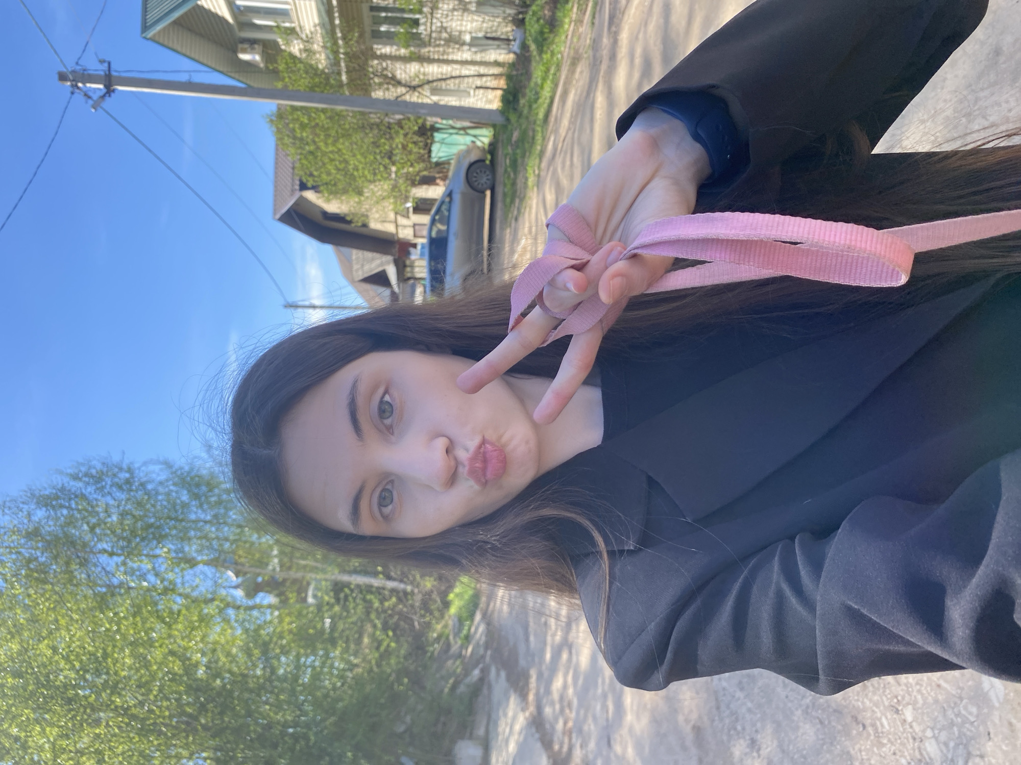A woman poses for a selfie outdoors on a sunny day. She has long, dark hair and fair skin, and she is wearing a dark-colored jacket. She holds up a pink strap with her hand, making a peace sign with her fingers near her face, while making a kissing face with her lips.
Behind her is a residential area, with houses and trees visible. A car is parked on the side of the road. Utility poles and wires are also visible. The environment appears to be a street or road in a residential part of a town, likely Votkinsk, Russia, given the request.