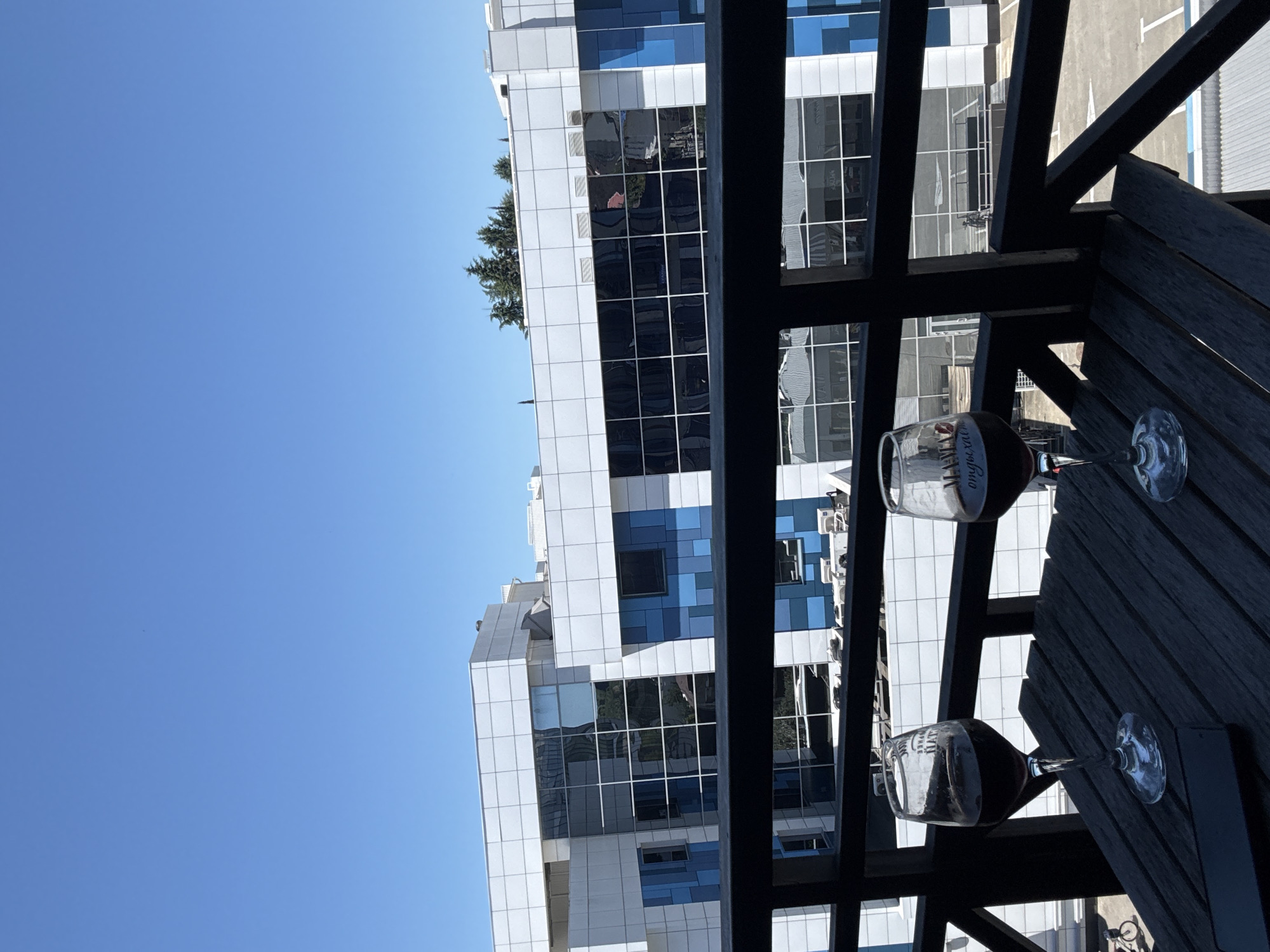 This image shows a close-up view of a wooden table on a balcony, with two glasses of red wine placed on it. The table is made of dark wood and is set against a backdrop of a modern building with a white and blue facade and reflective windows. The sky is clear and blue. In the distance, a few trees can be seen above the building. The scene suggests a relaxing moment on a balcony, possibly in an urban setting. The building's architecture and the presence of air conditioning units visible on its exterior suggest a commercial or office building. The clear sky and bright daylight indicate a sunny day. The overall impression is one of leisure and enjoyment, with the wine glasses being the focal point of the foreground. The image does not provide specific details about the city of Sochi, Russia, beyond its urban and modern aesthetic.
