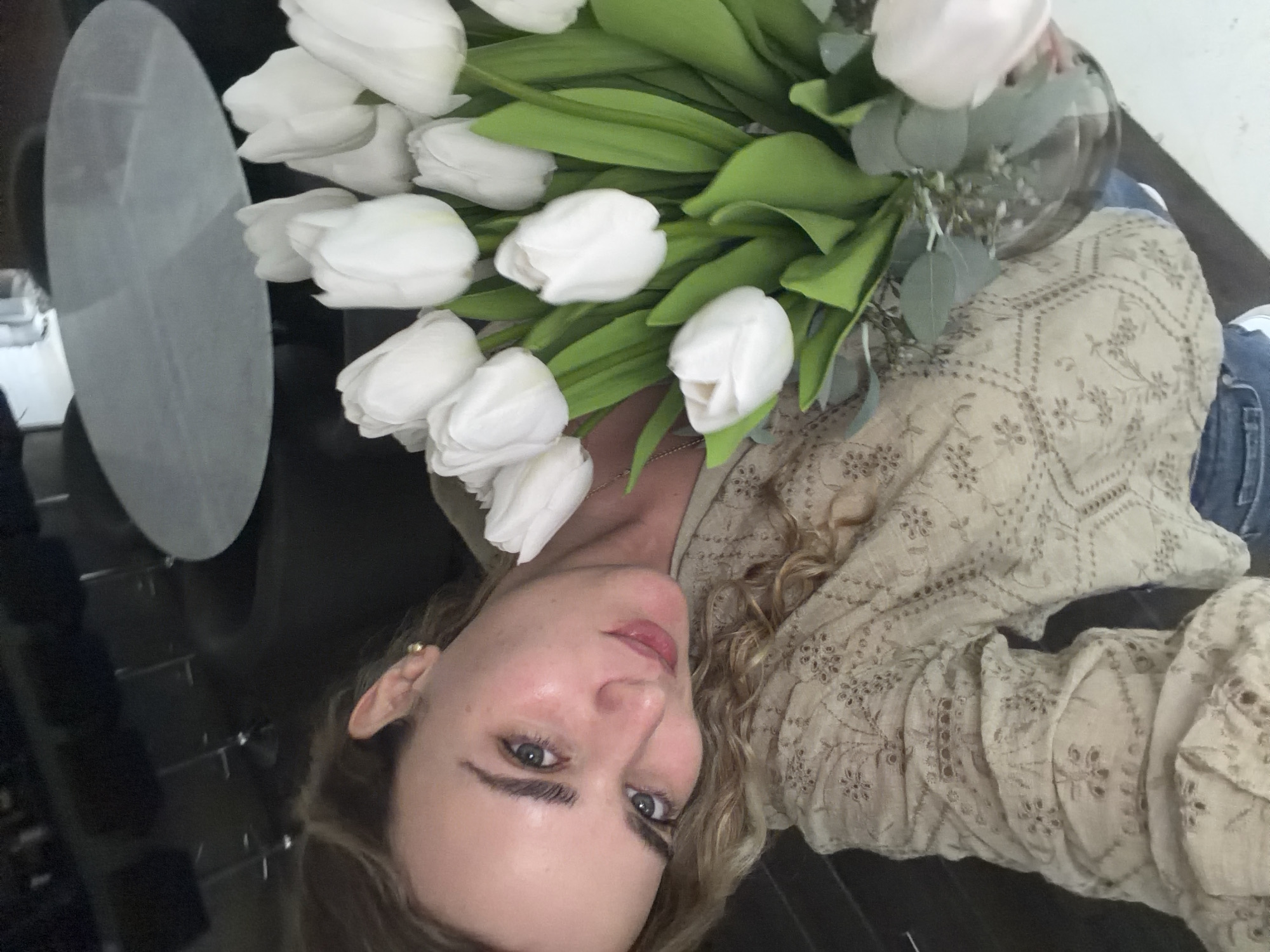 A woman is holding a bouquet of white tulips in front of her face. She is wearing a light brown embroidered shirt and blue jeans. She is sitting on a black surface, likely a table or countertop, and there is a gray oval object to her left. The location is unknown and there is no information in the image to support San Bernardino Tlaxcalancingo, Mexico.